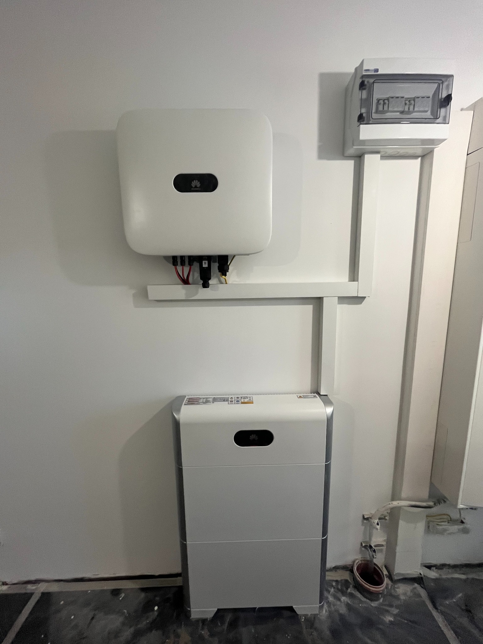 Biała instalacja fotowoltaiczna Huawei na ścianie, z akumulatorem i skrzynką elektryczną. Widoczne przewody i elementy montażowe. Minimalistyczny design.