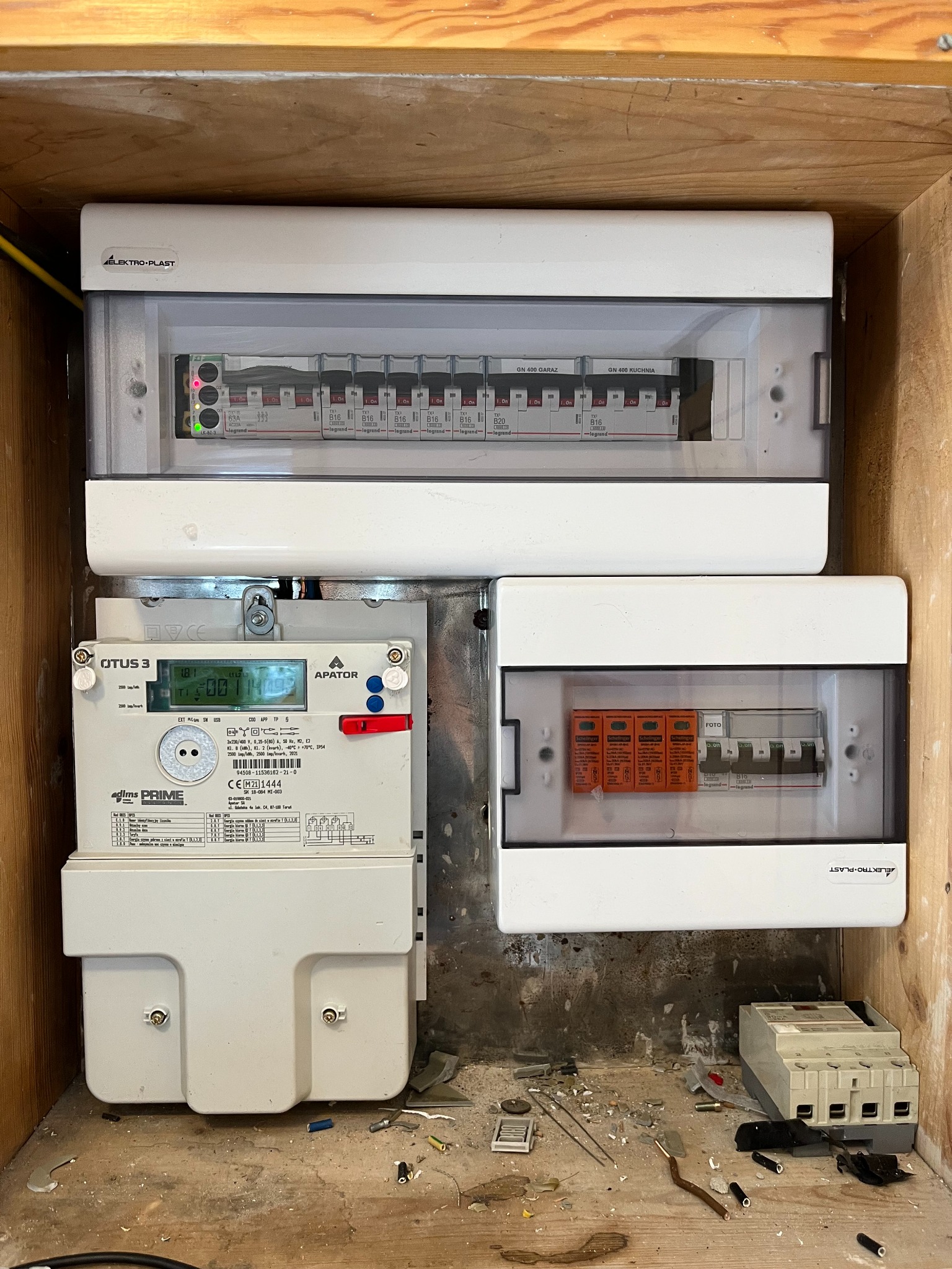 Rozdzielnia elektryczna z licznikiem energii i zabezpieczeniami w drewnianej obudowie. Widoczne bezpieczniki, pomiar energii i kable.