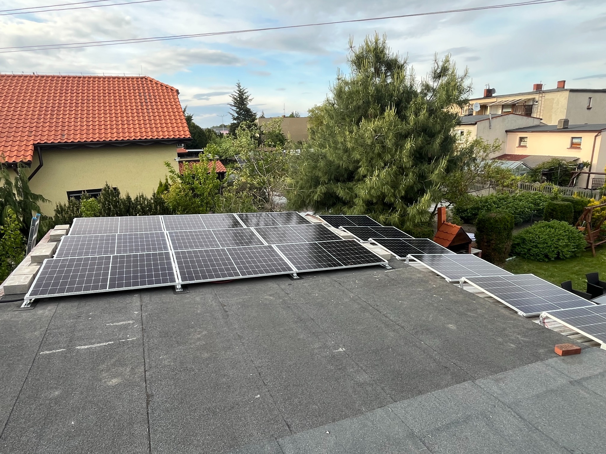 Montaż paneli słonecznych na dachu domu jednorodzinnego. Panele ułożone rzędami, widok z góry na instalację w trakcie realizacji. W tle zabudowa mieszkalna i zieleń.