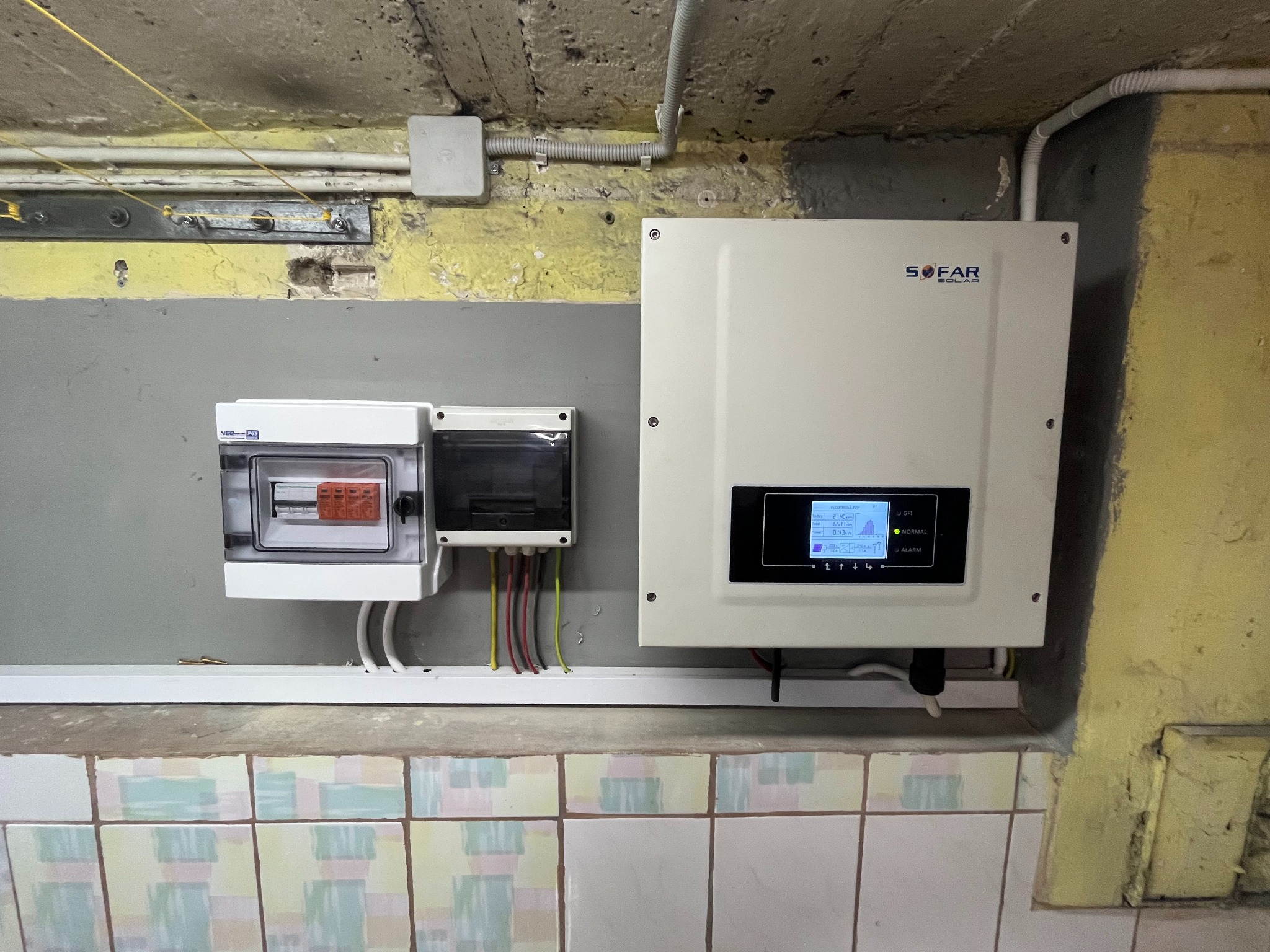 Zamontowany inwerter Sofar Solar obok skrzynki elektrycznej z bezpiecznikami, widoczne przewody i rury instalacyjne na tle ściany z płytkami i surowego betonu.