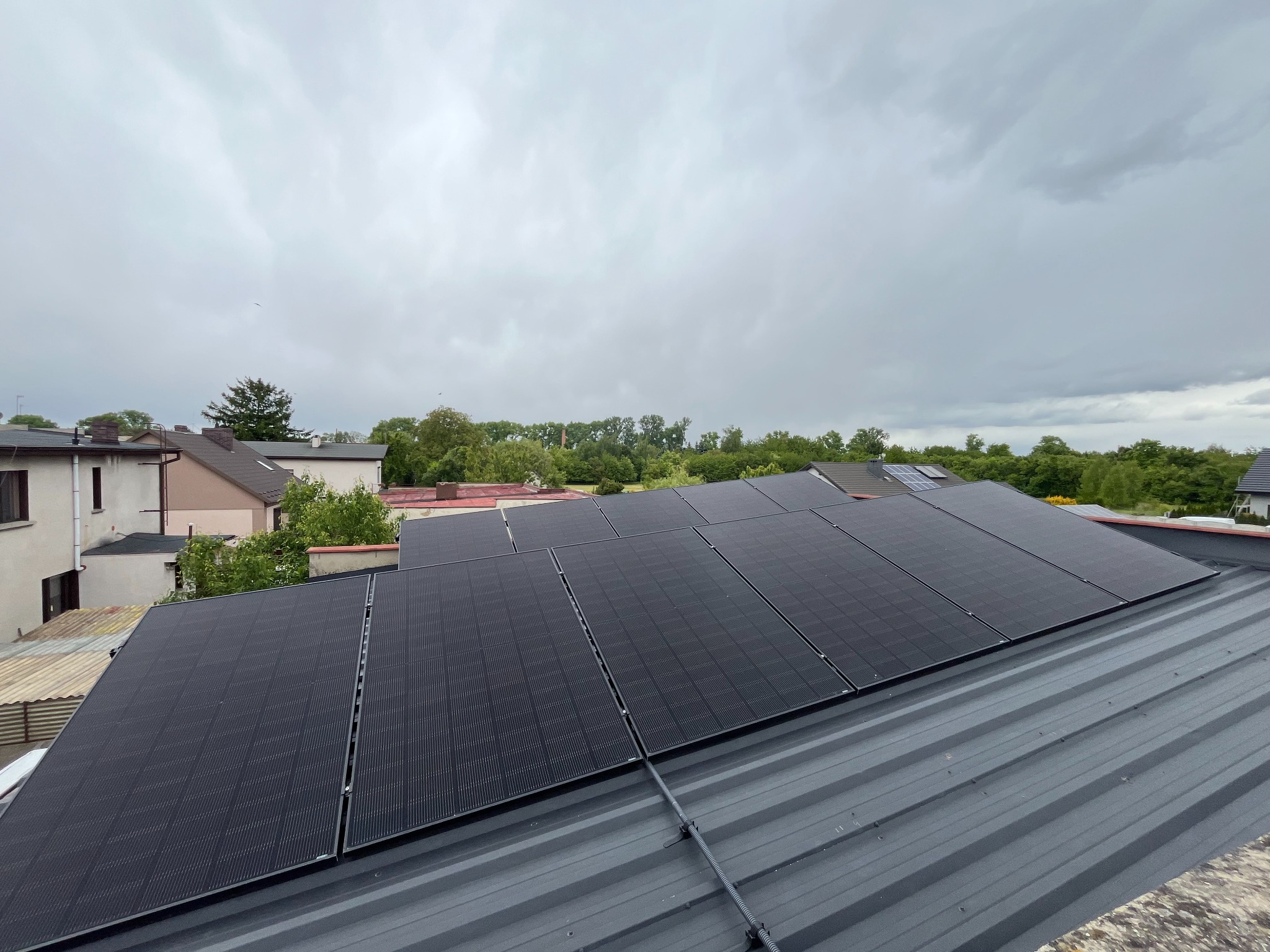 Panele fotowoltaiczne na szarym dachu blaszanej konstrukcji, widok z góry na tle zabudowań i pochmurnego nieba. Widoczne mocowania paneli.