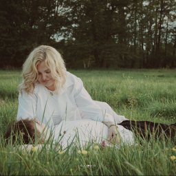 CloudPhotography - Kobieta z blond włosami w białej koszuli patrzy na leżącą osobę w trawie, w tle drzewa. Romantyczna sesja w plenerze, delikatne światło.