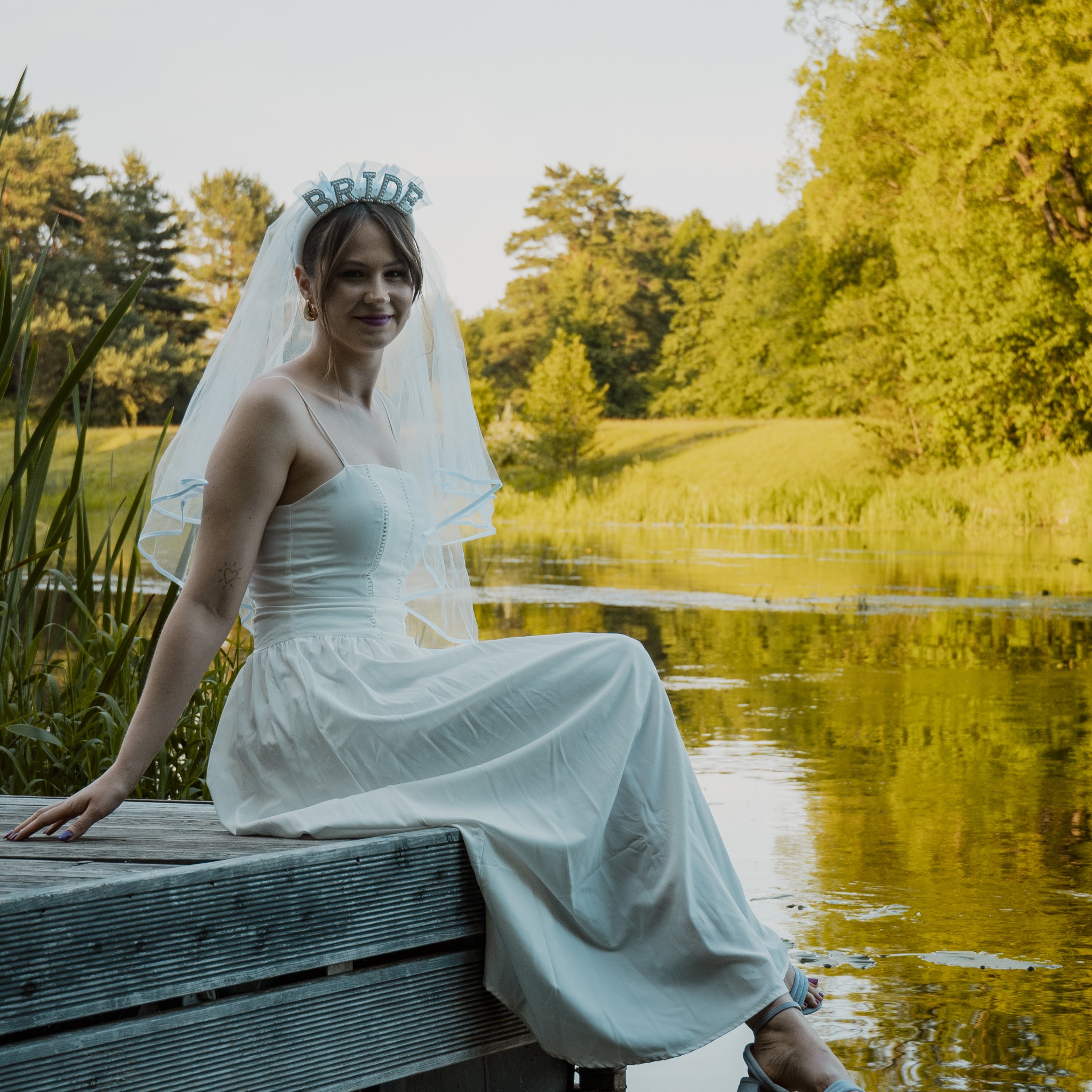 Panna młoda w zwiewnej sukni siedzi na drewnianym pomoście nad jeziorem, w tle zieleń drzew. Na głowie ma welon i diadem z napisem 'Bride'. Romantyczna sesja plenerowa.