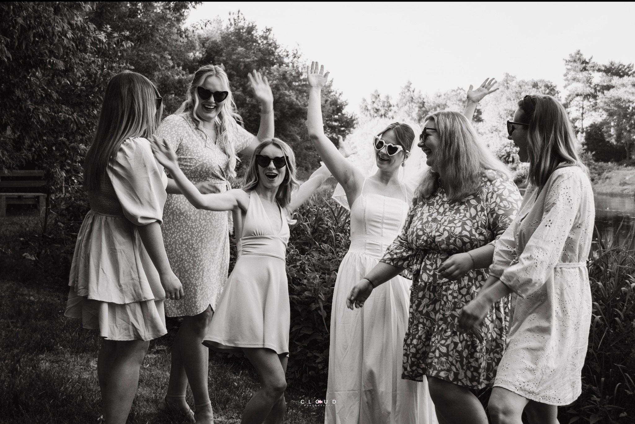 Czarno-białe zdjęcie grupy roześmianych kobiet w okularach przeciwsłonecznych, unoszących ręce w górę, na tle zieleni. Jedna z nich ma welon.