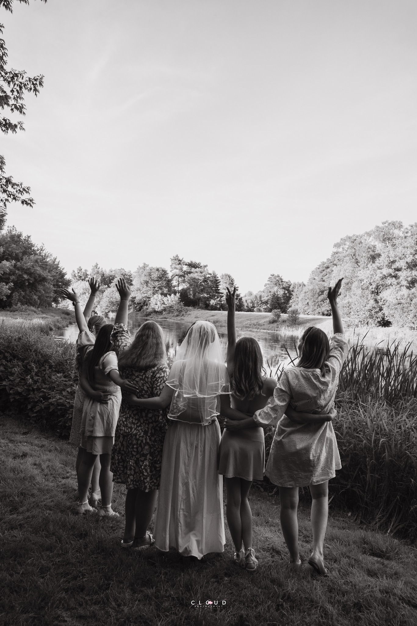 Czarno-białe zdjęcie panny młodej z druhnami, stojących tyłem i trzymających ręce w górze, na tle jeziora i drzew. Ujęcie z perspektywy ziemi.
