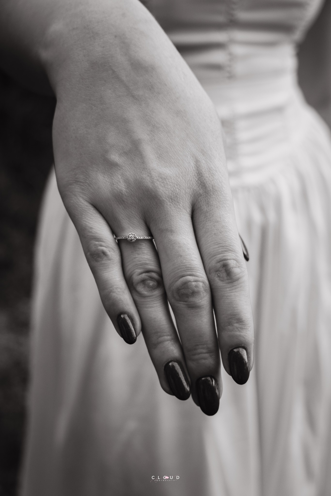 Czarno-białe zbliżenie dłoni kobiety z pierścionkiem zaręczynowym na tle sukni ślubnej, ukazujące detale biżuterii i manicure.