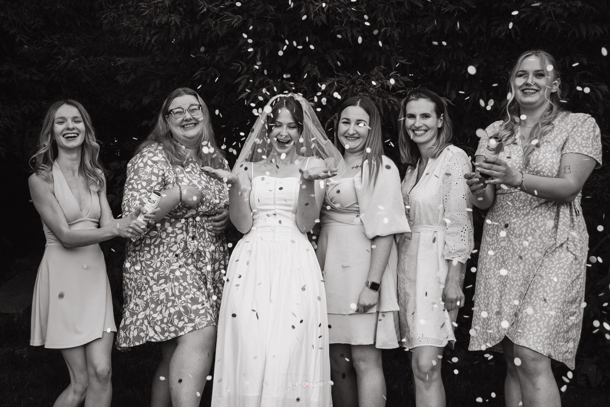 Czarno-białe zdjęcie panny młodej w welonie i sukni otoczonej przez druhny rzucające konfetti na tle zieleni, radosna atmosfera wieczoru panieńskiego.