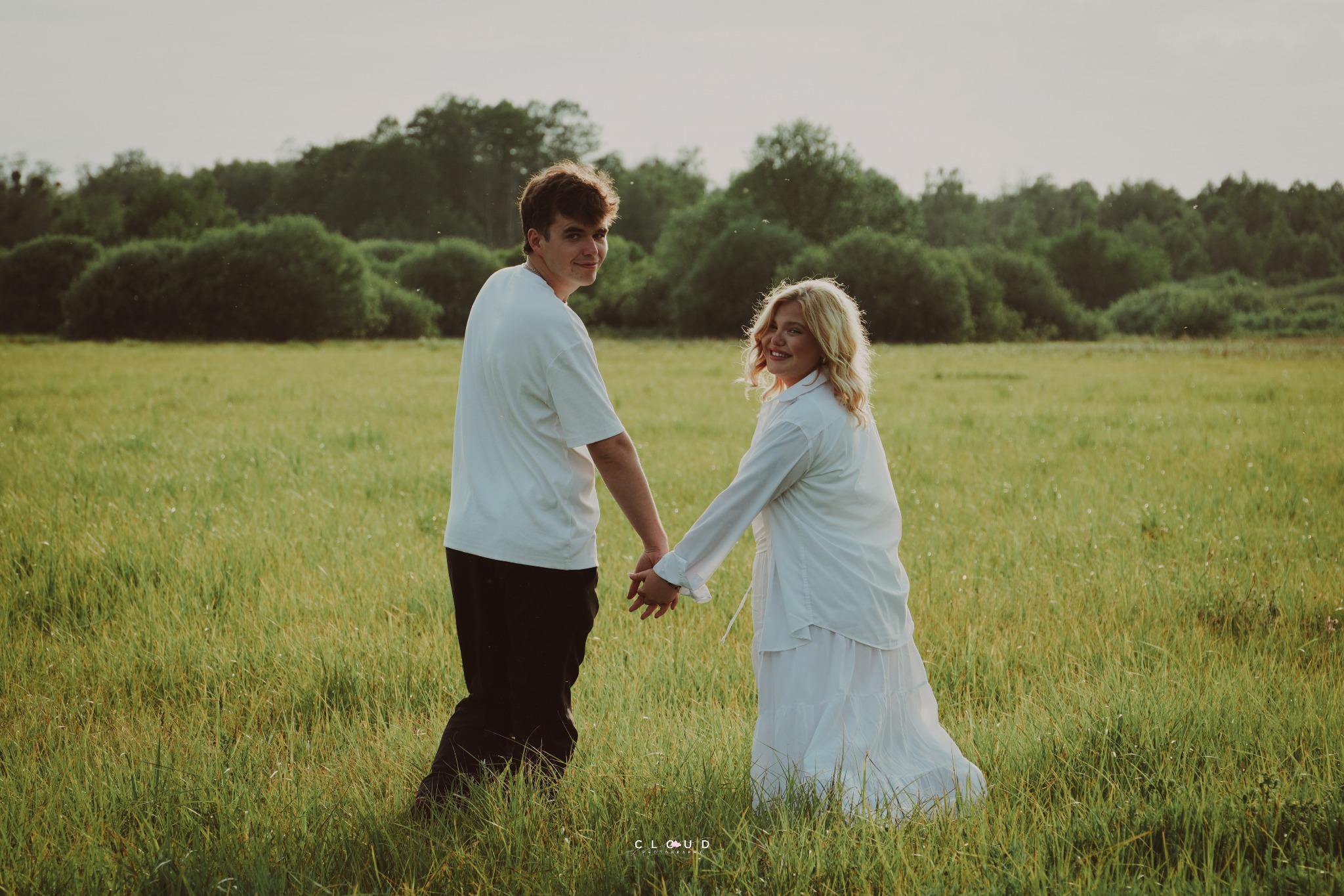 Zakochana para trzyma się za ręce na łące, oboje ubrani na biało. Mężczyzna spogląda w obiektyw, a kobieta uśmiecha się, patrząc w jego stronę. Romantyczna sesja w plenerze.