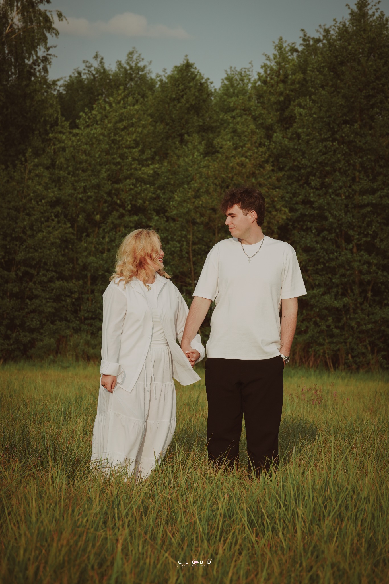 Zakochana para trzymająca się za ręce na łące w Płońsku, w tle zielony las. Romantyczna sesja narzeczeńska w naturalnym otoczeniu, ciepłe światło.