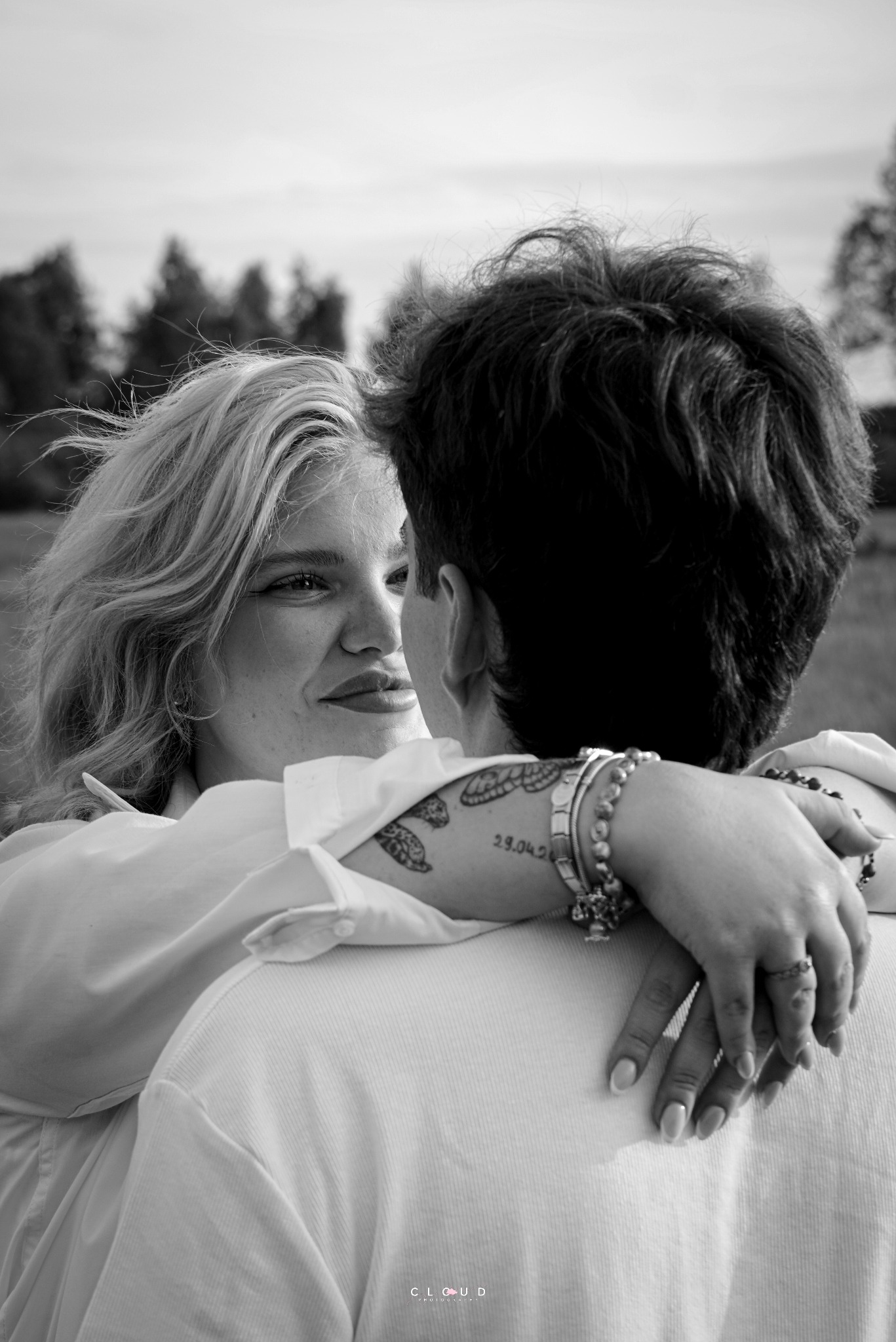 Czarno-białe zbliżenie na uśmiechniętą blondynkę obejmującą partnera z widocznym tatuażem na ramieniu i bransoletkami, na tle łąki i drzew.