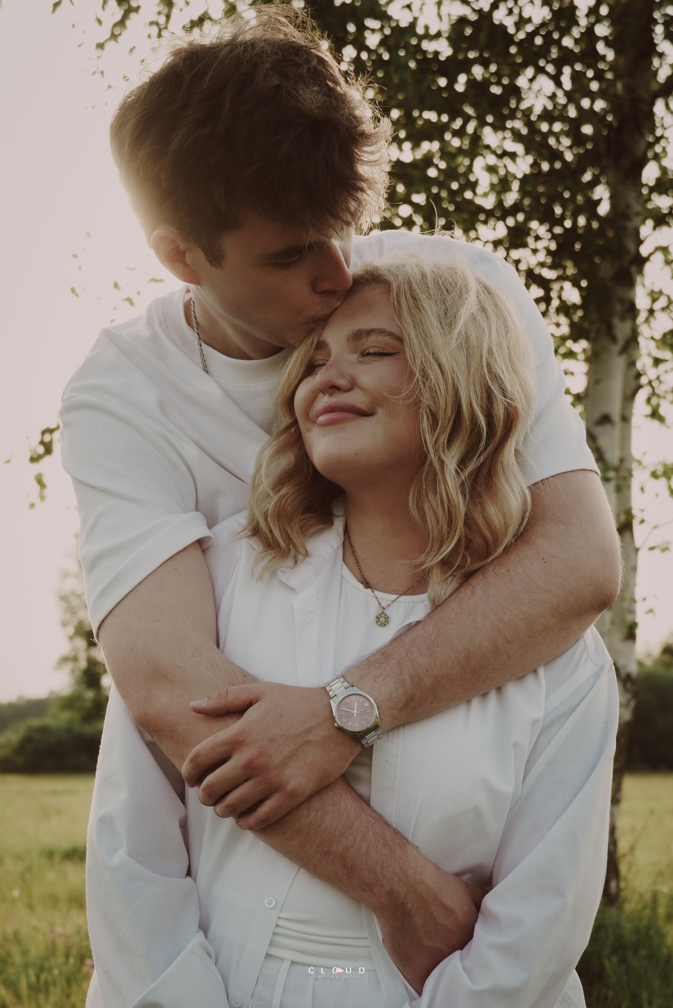 Młody mężczyzna czule całuje w czoło uśmiechniętą blondynkę na tle zieleni. Para ubrana w białe stroje, subtelne światło słoneczne. Romantyczna sesja plenerowa.