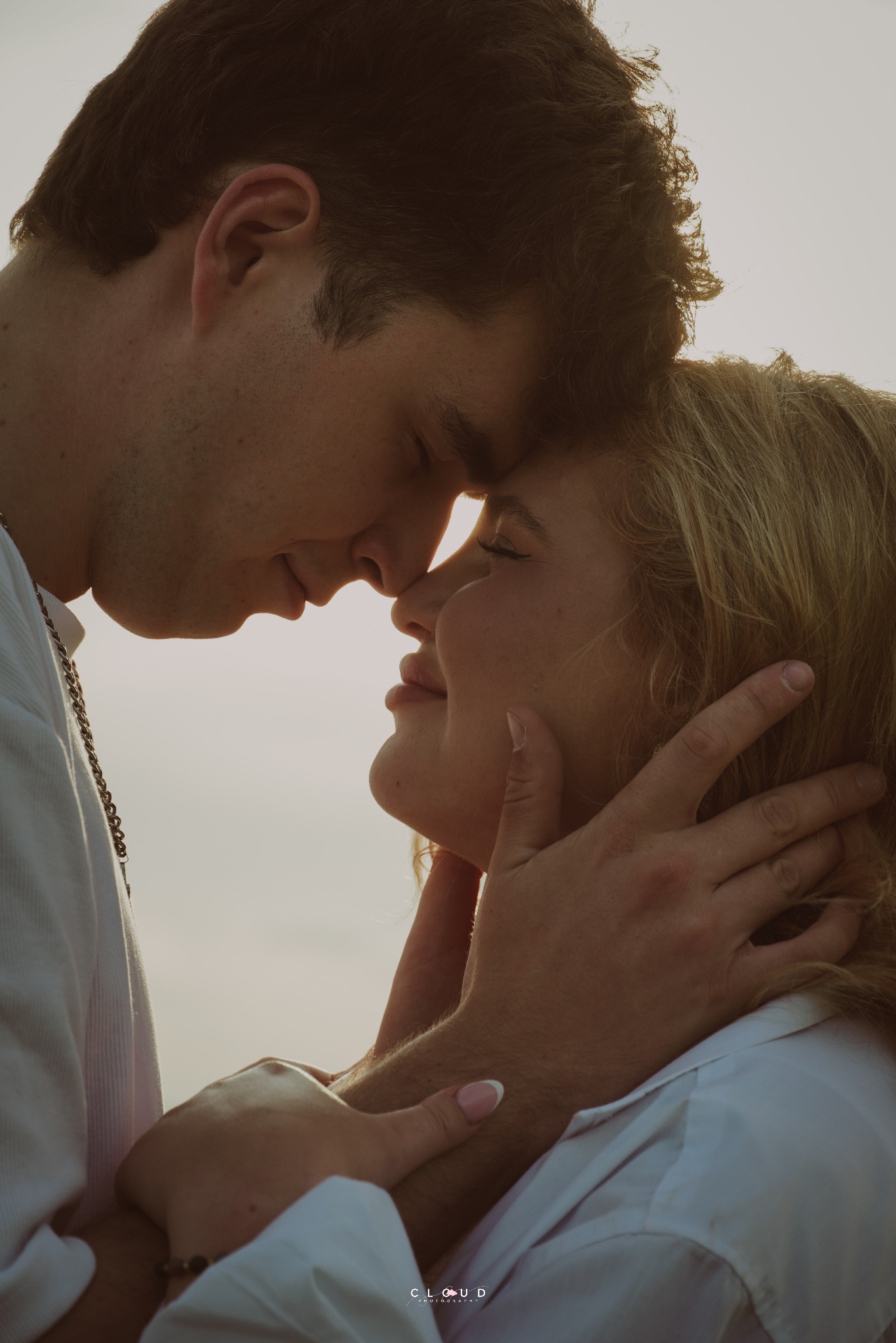 Zakochana para w zbliżeniu, dotykająca się nosami w miękkim, ciepłym świetle. On w białej koszuli i łańcuszku, ona z blond włosami, delikatnie obejmowana.