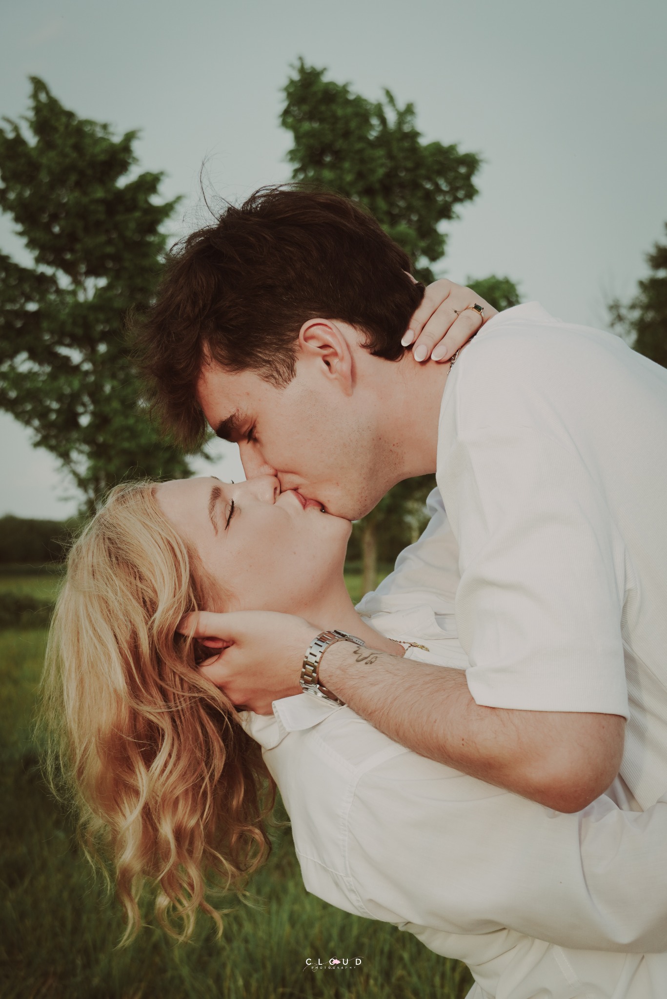 Romantyczne ujęcie: całująca się para na tle zieleni. Kobieta z blond lokami i mężczyzna w białej koszuli, czuły moment uchwycony z bliska, z dbałością o detale.