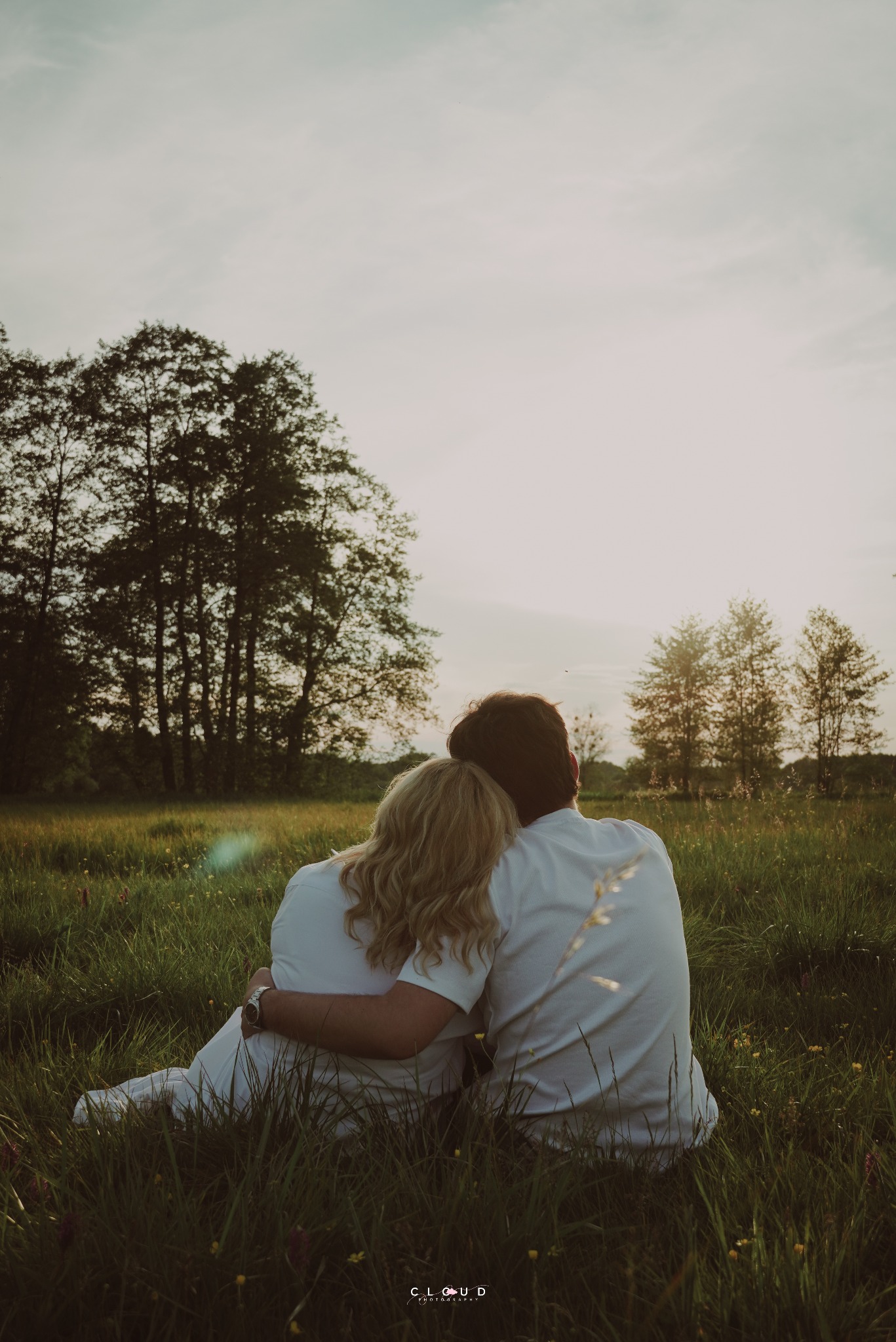Para w białych koszulach siedzi w trawie, obejmując się i patrząc w dal, w tle drzewa i zachodzące słońce. Romantyczna sesja w plenerze.