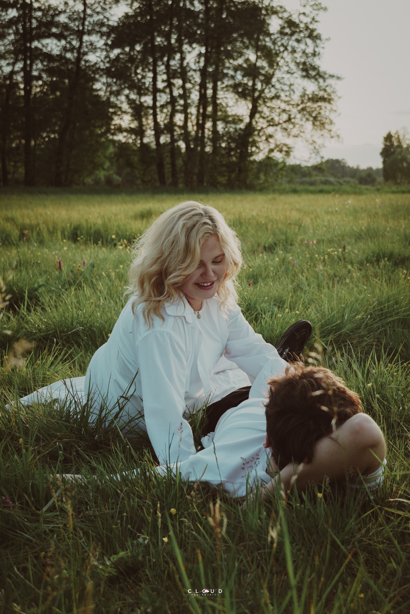 Romantyczna sesja w plenerze: uśmiechnięta blondynka w białej koszuli siedzi na trawie obok leżącego bruneta. W tle drzewa i łąka w ciepłym świetle.