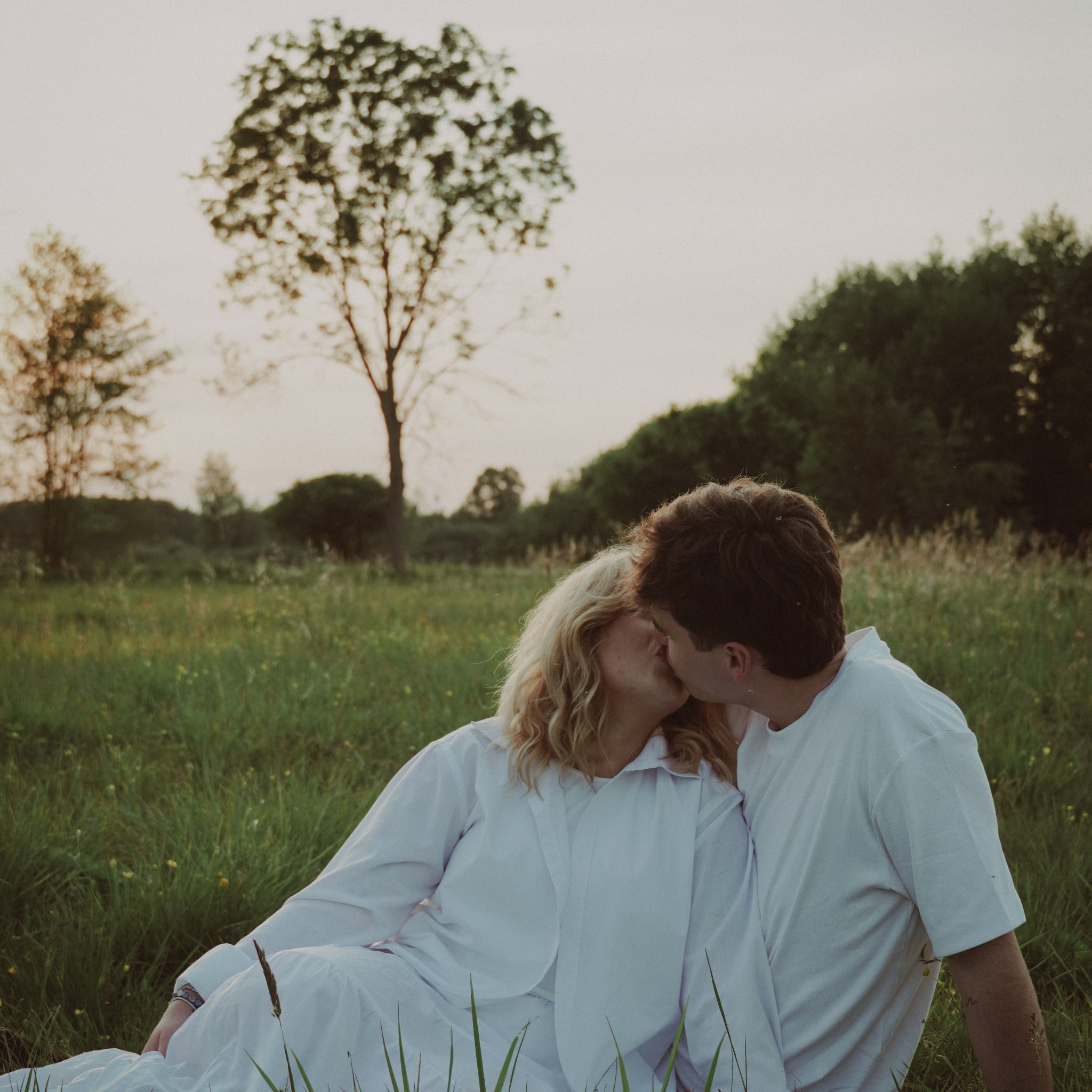 Zakochana para w białych ubraniach całuje się na łące o zmierzchu. Romantyczna sesja w plenerze, wysoka trawa, drzewa w tle, stonowana kolorystyka.