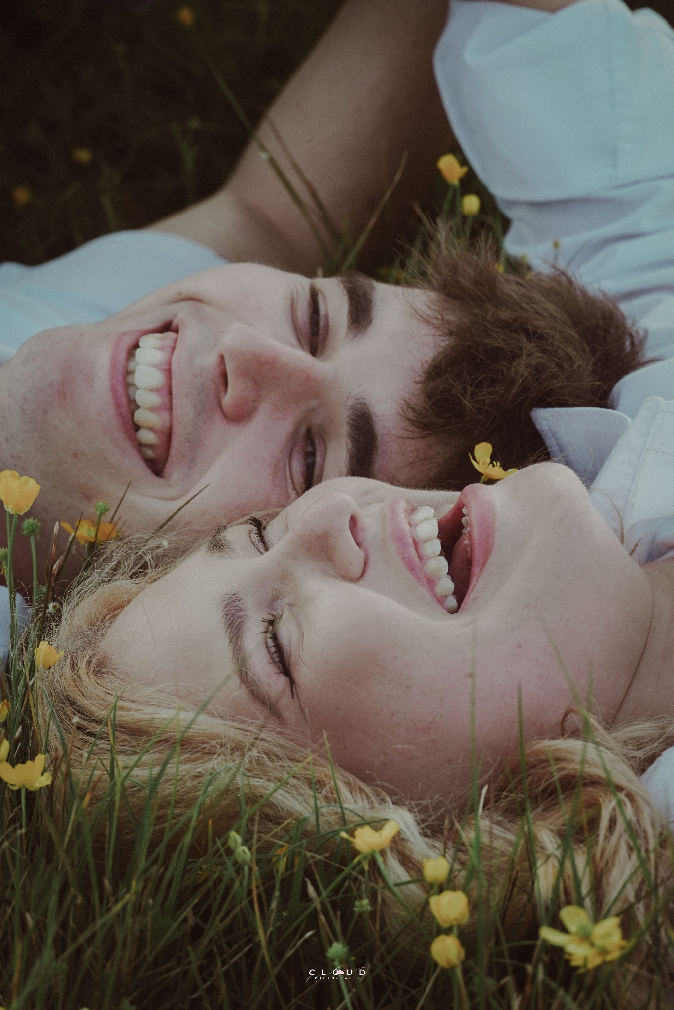 Uśmiechnięta para leży w trawie wśród żółtych kwiatów, zbliżenie na twarze. Mężczyzna z ciemnymi włosami i kobieta z blond włosami, oboje ubrani w białe koszule.