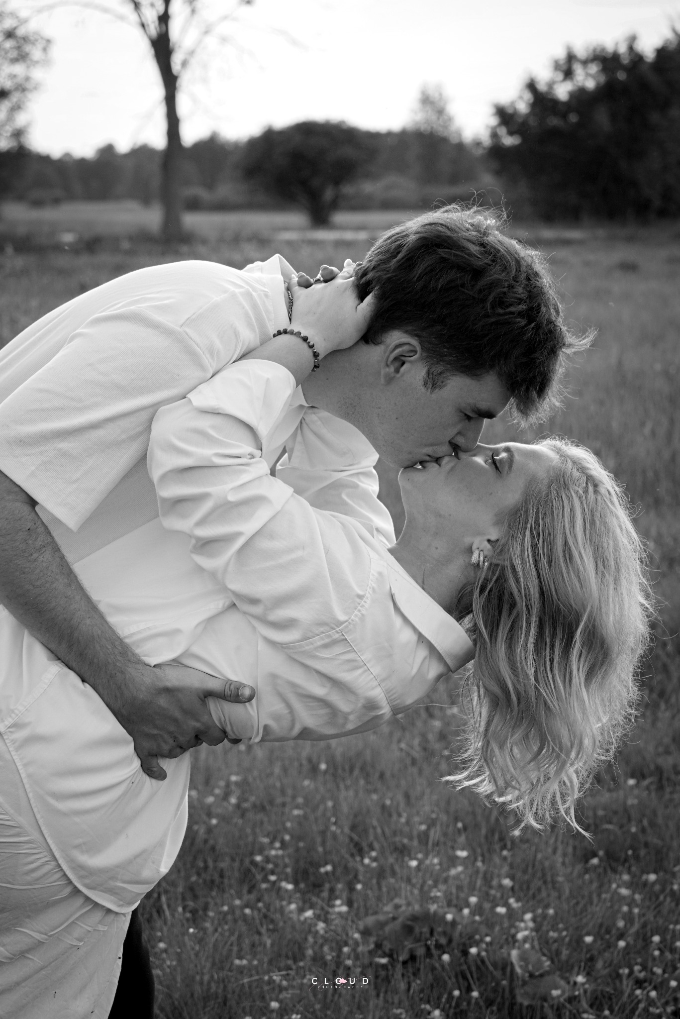 Czarno-biała fotografia narzeczeńska: para całująca się na łące, on obejmuje ją w pasie, a ona odchyla się do tyłu. Romantyczny kadr w naturalnym otoczeniu.