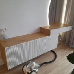 Nowoczesna toaletka z oświetleniem LED i ryflowanymi frontami, z blatem w odcieniu drewna. Na blacie płyn do czyszczenia. Obok odkurzacz.