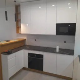 Nowoczesna kuchnia z białymi frontami, czarnym blatem i wbudowanym piekarnikiem. Widoczne oświetlenie punktowe i drewniane elementy.