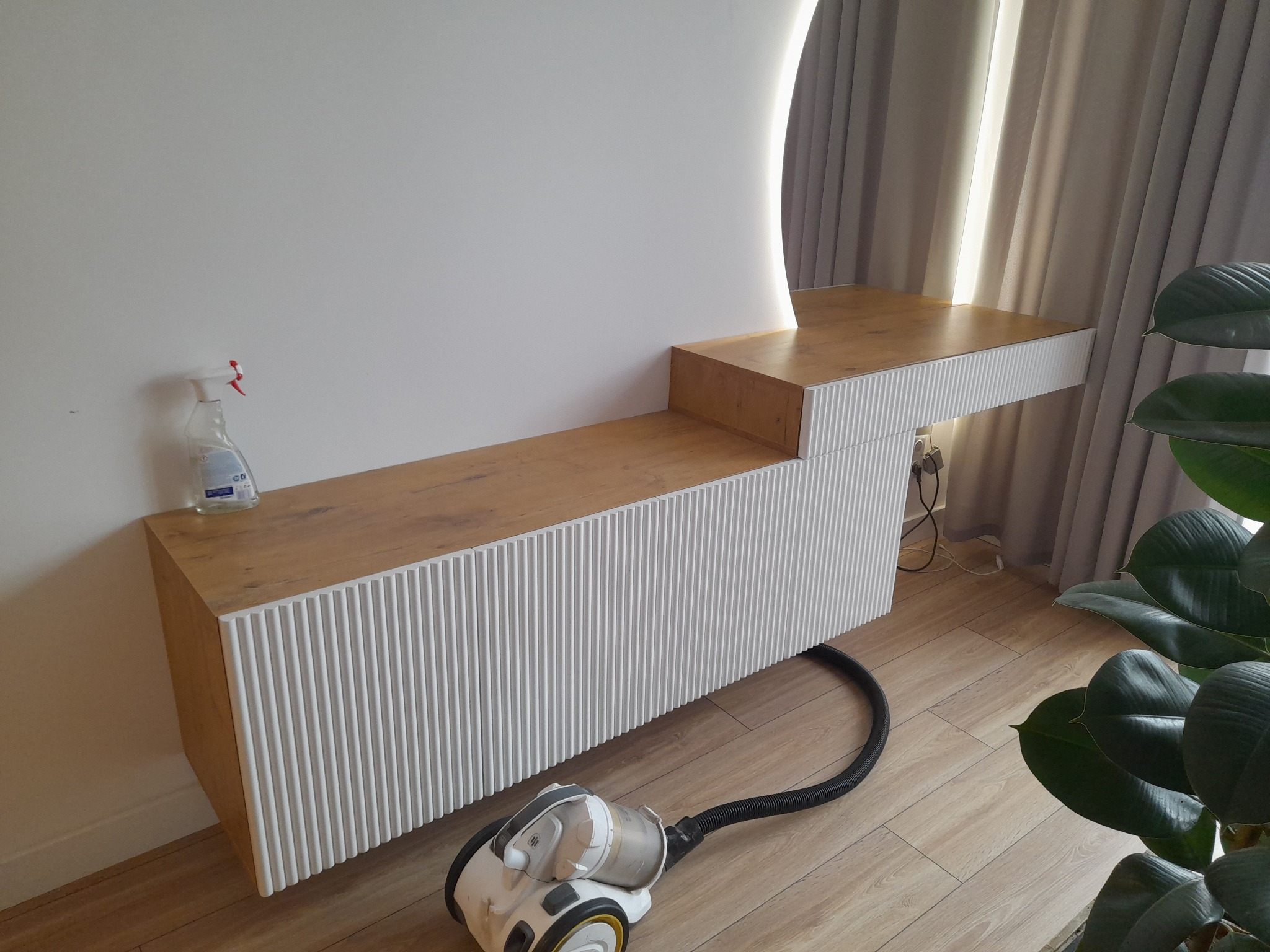 Nowoczesna toaletka z oświetleniem LED i ryflowanymi frontami, z blatem w odcieniu drewna. Na blacie płyn do czyszczenia. Obok odkurzacz.