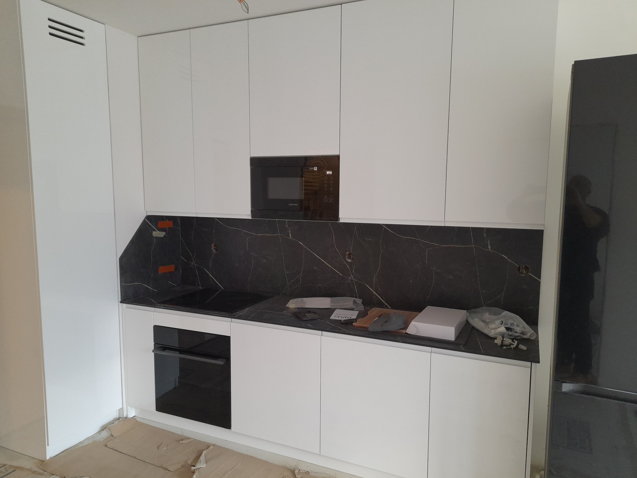 Biała, nowoczesna kuchnia z czarnym blatem i zabudowaną mikrofalówką. Widoczne akcesoria montażowe na blacie. Minimalistyczny design, proste linie.