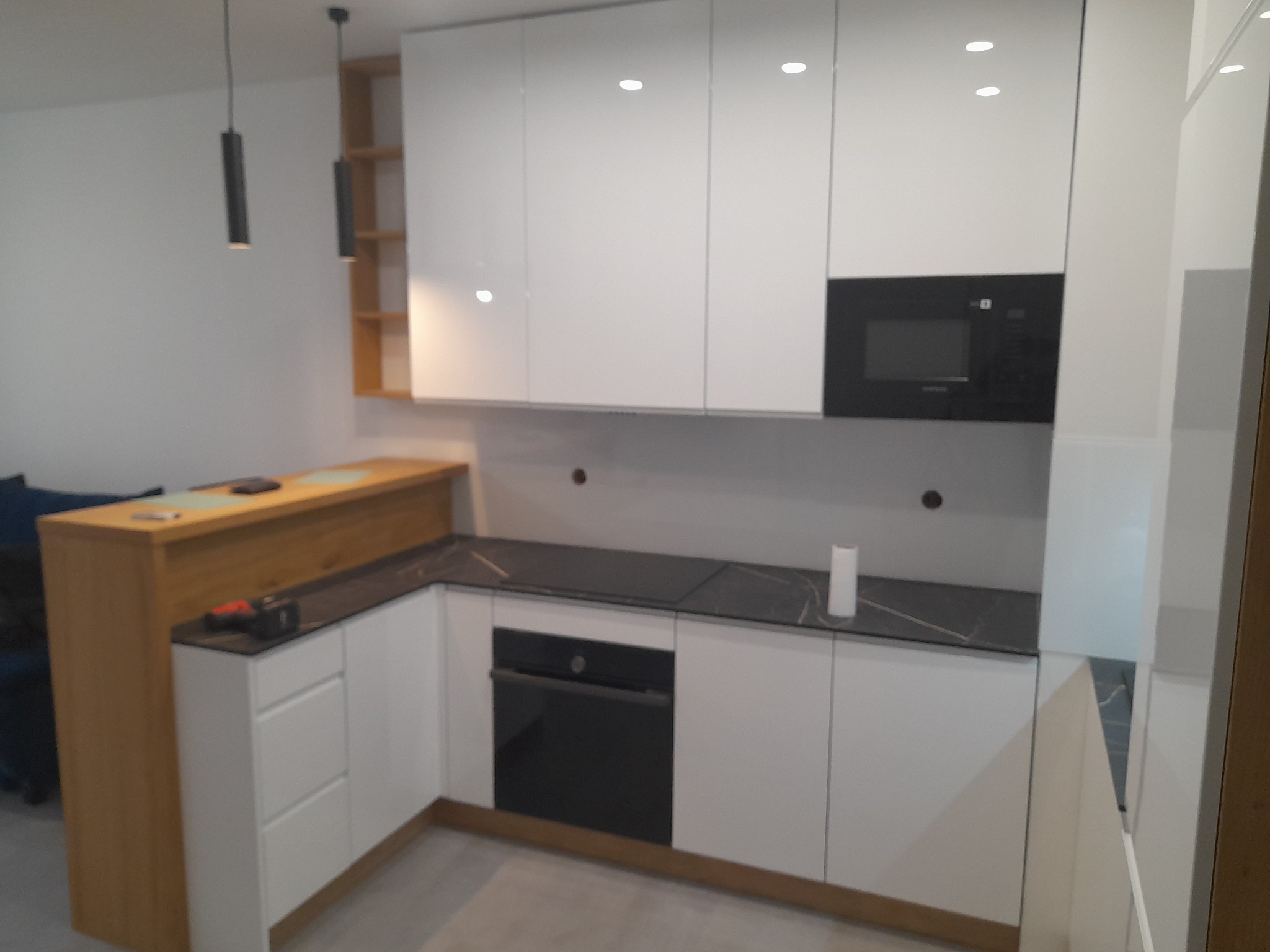 Nowoczesna kuchnia z białymi frontami, czarnym blatem i wbudowanym piekarnikiem. Widoczne oświetlenie punktowe i drewniane elementy.