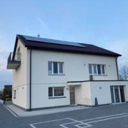 Nowoczesny dom z kremową elewacją i panelami słonecznymi na brązowym dachu. Widoczne okna, balkon i kostka brukowa na podjeździe. Ujęcie z dołu.