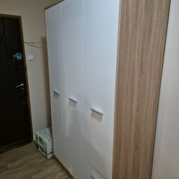 Stolarstwo Świerkowski - Nowoczesna szafa trzydrzwiowa z białymi frontami i drewnopodobnymi bokami, z szufladami na dole, stojąca w rogu pokoju obok drzwi.