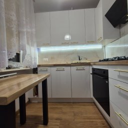 Stolarstwo Świerkowski - Nowoczesna kuchnia z białymi szafkami, złotymi uchwytami i drewnianym blatem. Oświetlenie LED pod szafkami górnymi. Widoczny fragment stołu z drewnianym blatem.