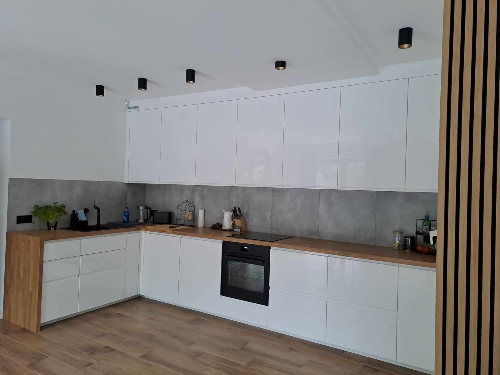 Nowoczesna kuchnia z białymi, lakierowanymi szafkami, drewnianym blatem i czarnymi lampami sufitowymi. Minimalistyczny design z drewnianą ścianką dekoracyjną.
