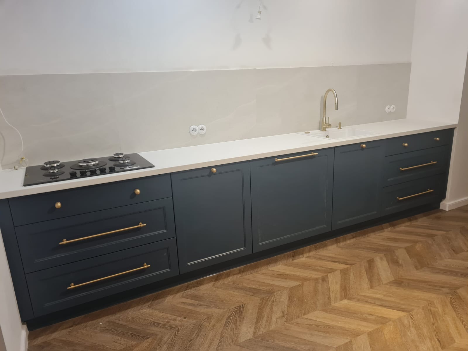 Granatowe szafki kuchenne z białym blatem i złotymi uchwytami, widok na zlew i płytę grzewczą. Parkiet ułożony w jodełkę.