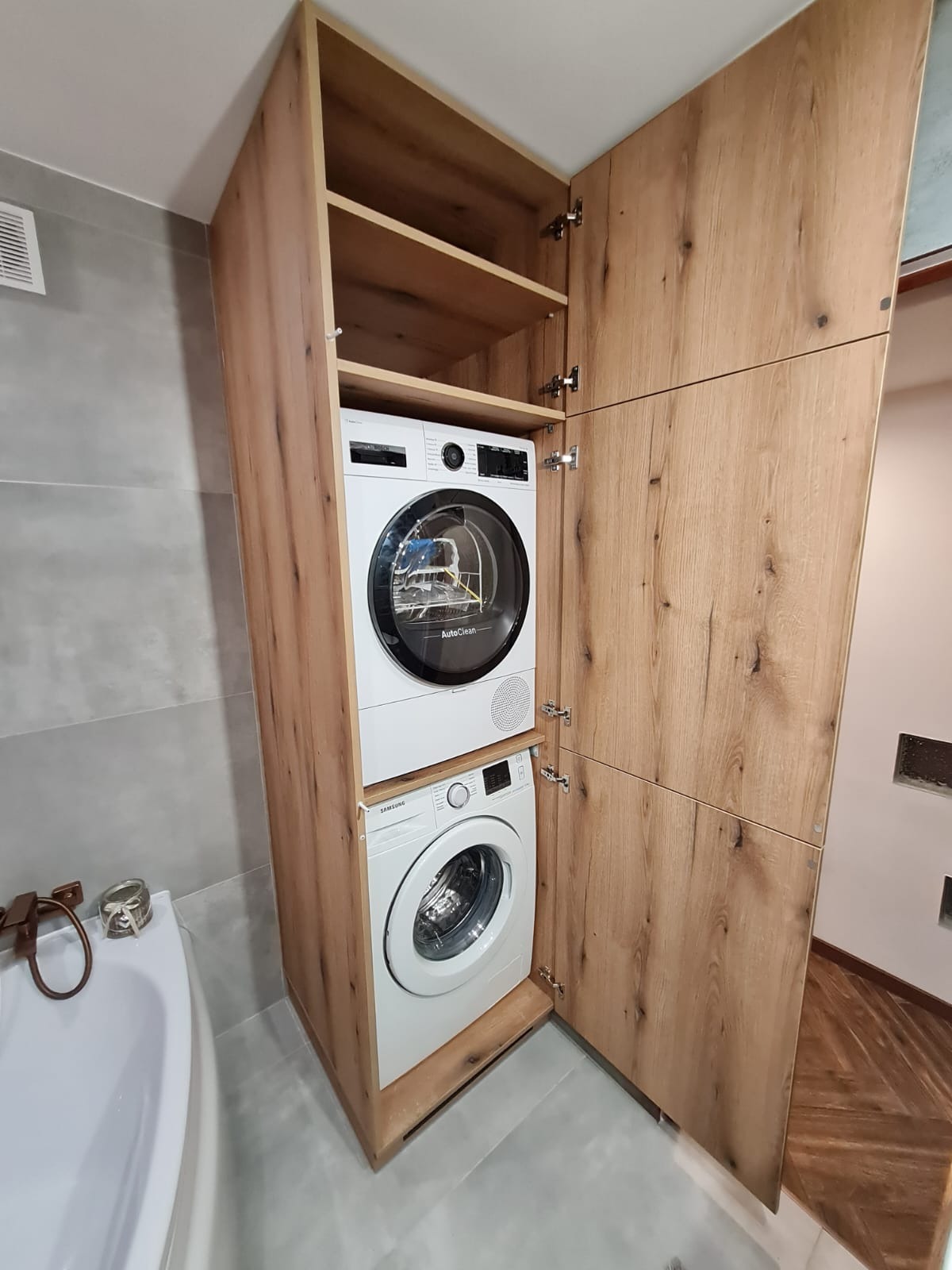 Biała pralka i suszarka w drewnianej zabudowie szafy w łazience. Otwarta szafa z półkami. Widoczne zawiasy i elementy montażowe. Ciekawy układ z drewnianym wykończeniem.
