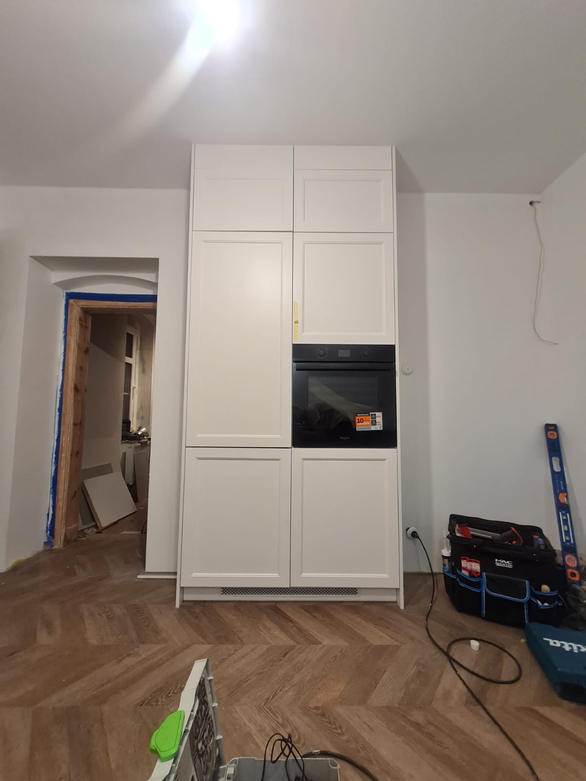 Biała zabudowa kuchenna z piekarnikiem w trakcie montażu, widoczny parkiet w jodełkę, narzędzia i poziomica. Drzwi do innego pomieszczenia z ościeżnicą w remoncie.