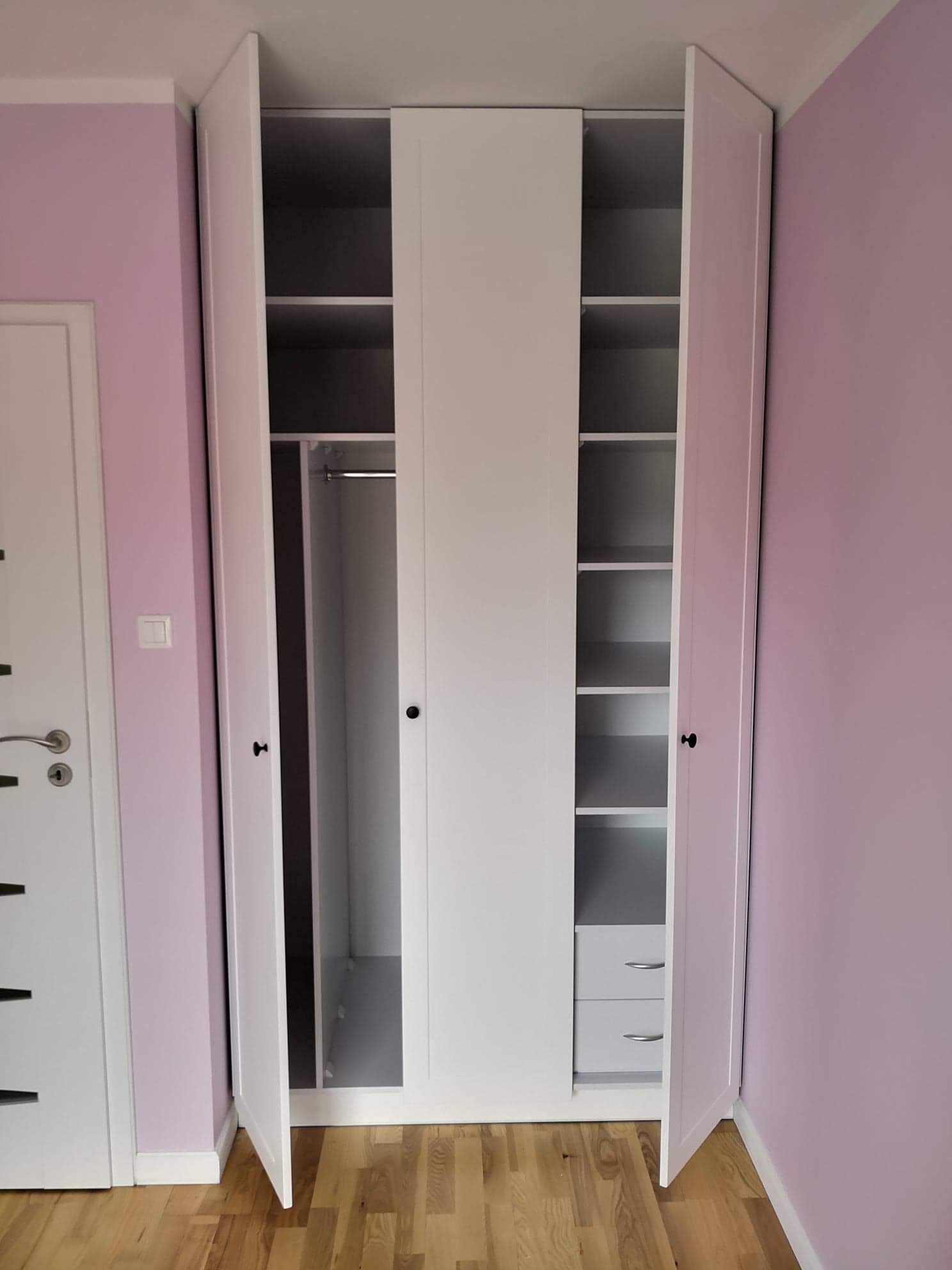 Biała, trzydrzwiowa szafa z otwartymi bocznymi drzwiami, ukazująca półki i drążek na ubrania, na tle fioletowej ściany i parkietu.