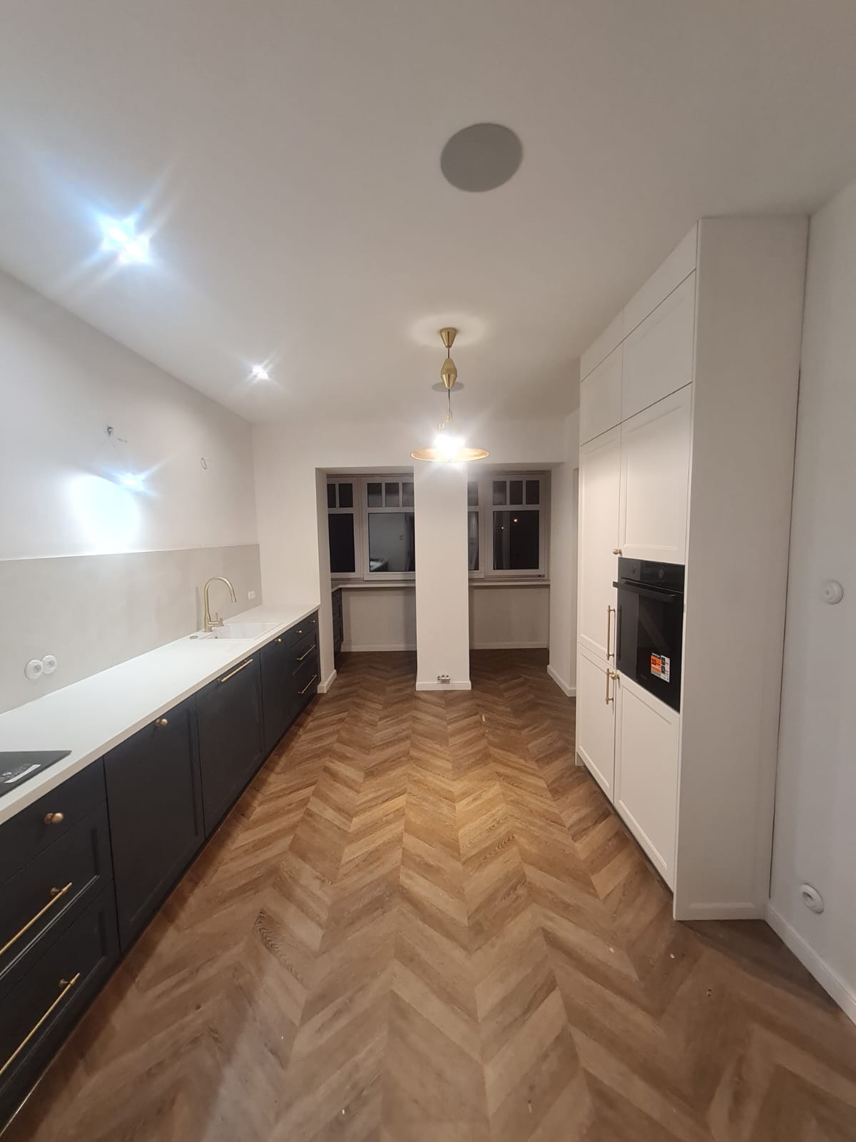 Nowoczesna kuchnia z ciemnymi szafkami, białym blatem, złotą armaturą i parkietem w jodełkę. Białe szafy z piekarnikiem w zabudowie. Lampa sufitowa w stylu retro.