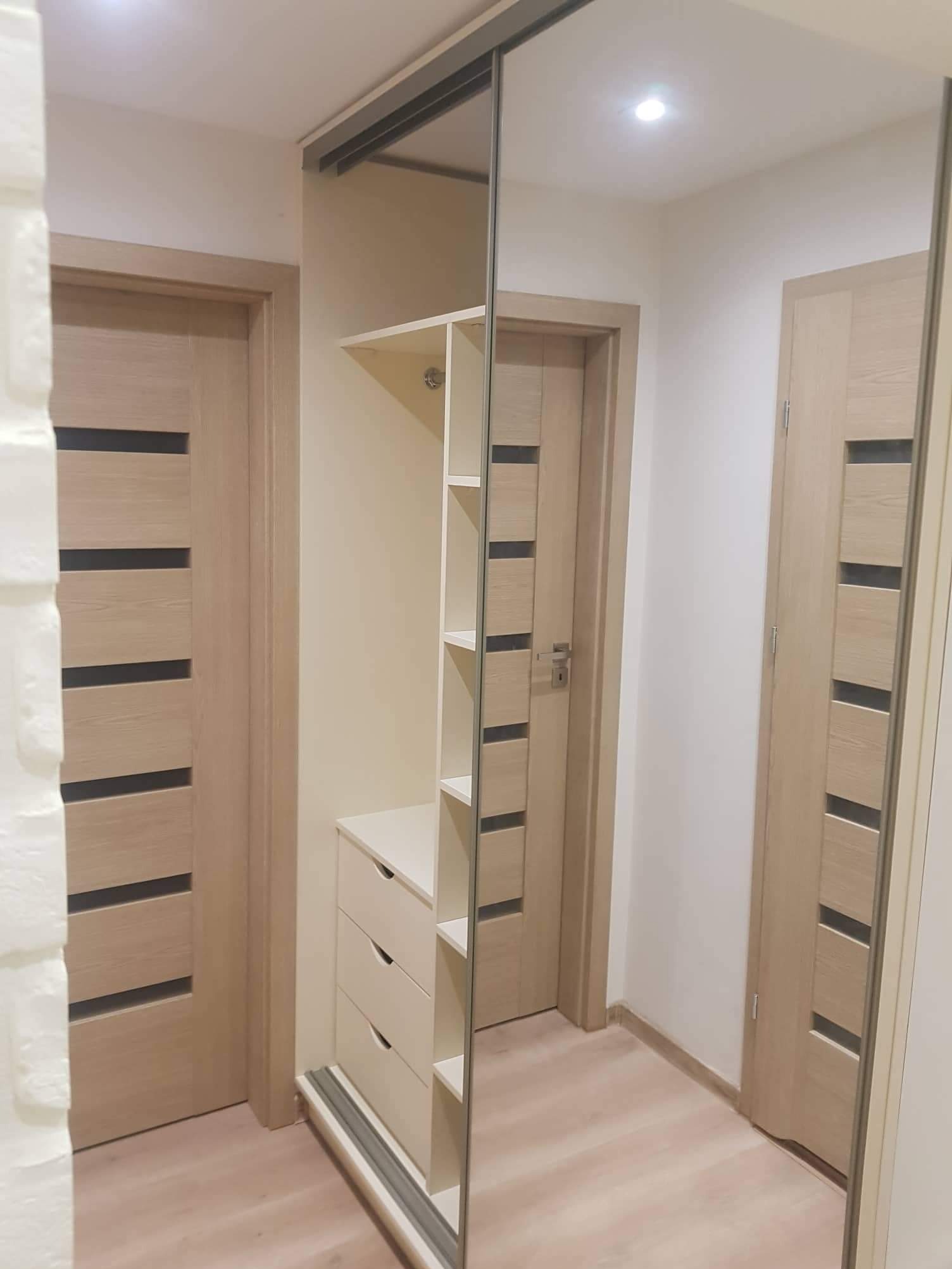 Beżowa szafa wnękowa z lustrzanymi drzwiami przesuwnymi, widoczna z perspektywy wnętrza, z dwiema parami drzwi po bokach. Jasne drewno, minimalistyczny design.