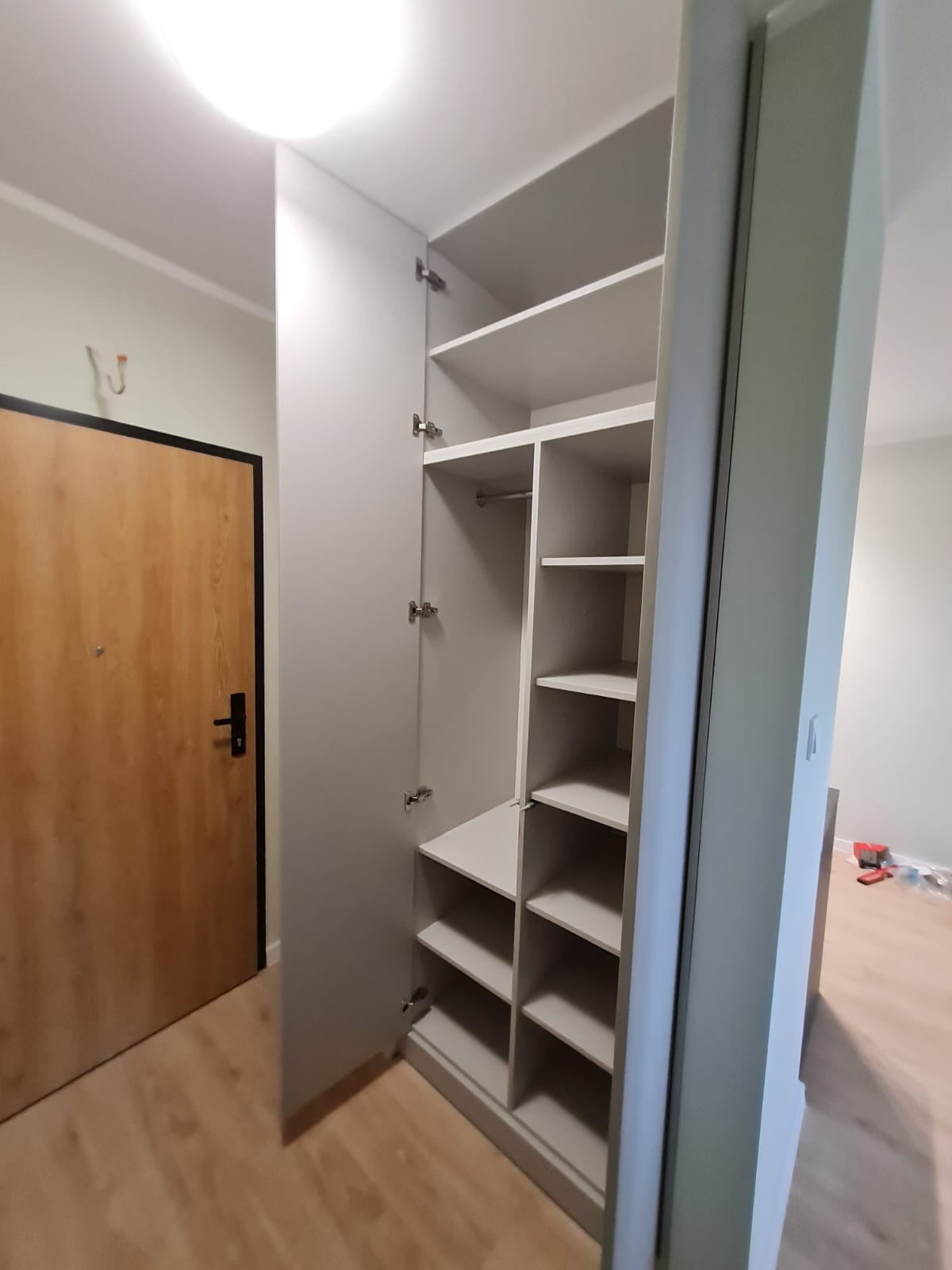 Wnętrze przedpokoju z otwartą, szarą szafą wnękową z półkami i drążkiem na ubrania. Widoczny fragment drzwi wejściowych i podłoga. Minimalistyczny styl.