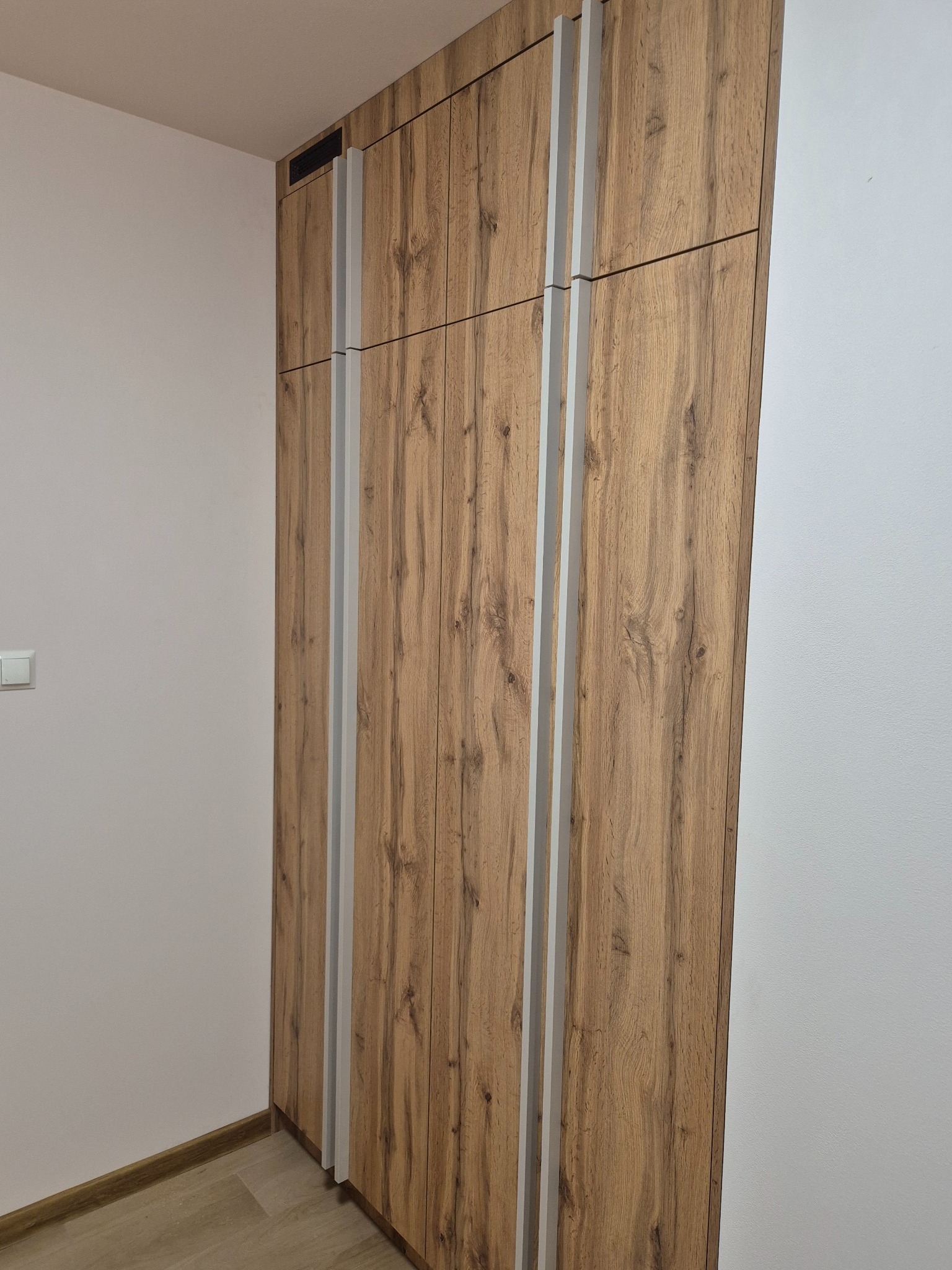 Wysoka, zabudowana szafa z drewnopodobnymi frontami i jasnymi, pionowymi listwami, umieszczona w rogu pomieszczenia z jasnymi ścianami i podłogą.