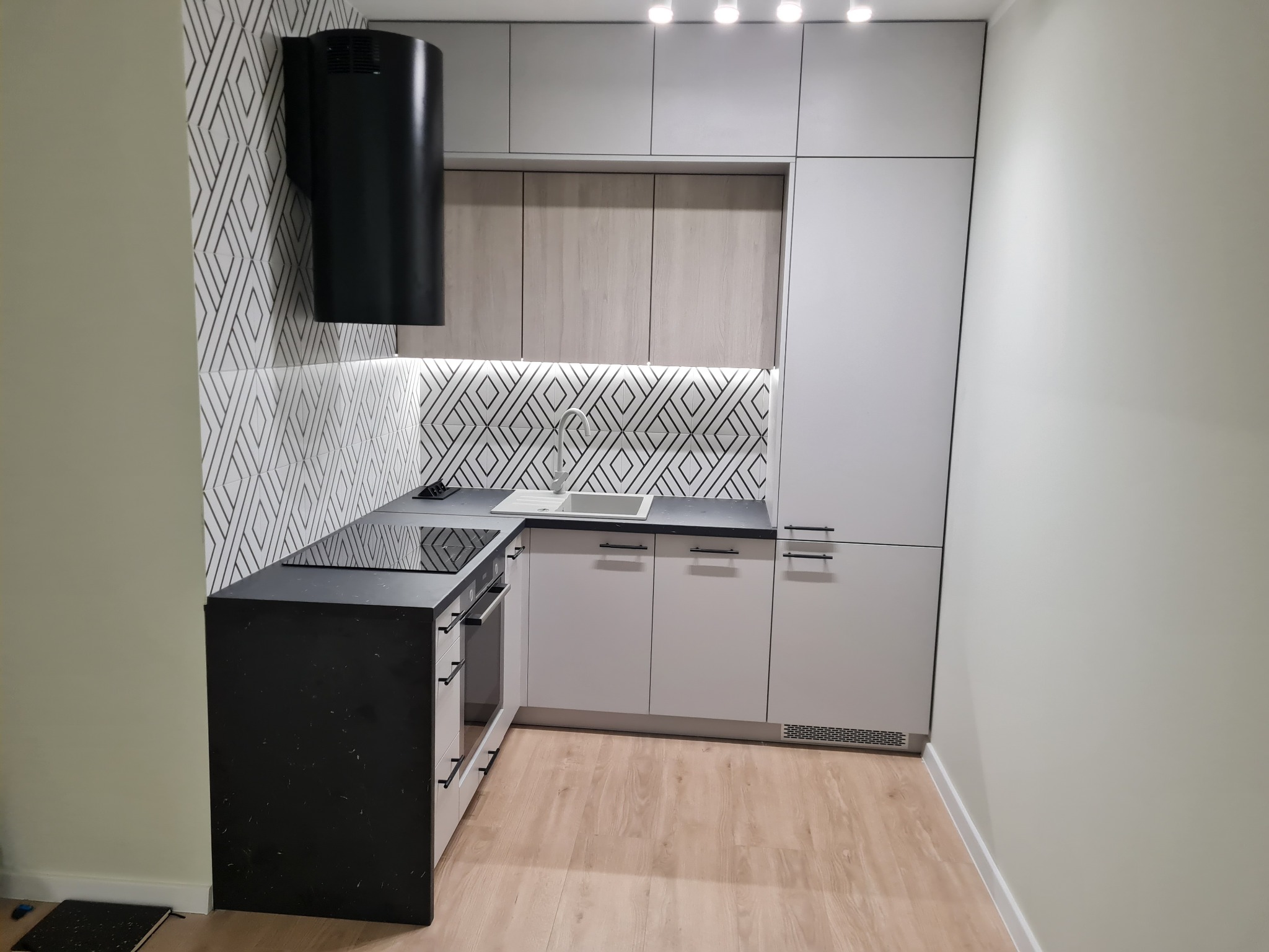 Nowoczesna, jasna kuchnia z czarnym blatem i okapem. Geometryczny wzór na ścianie, drewniane fronty szafek górnych. Minimalistyczny design, oświetlenie punktowe.