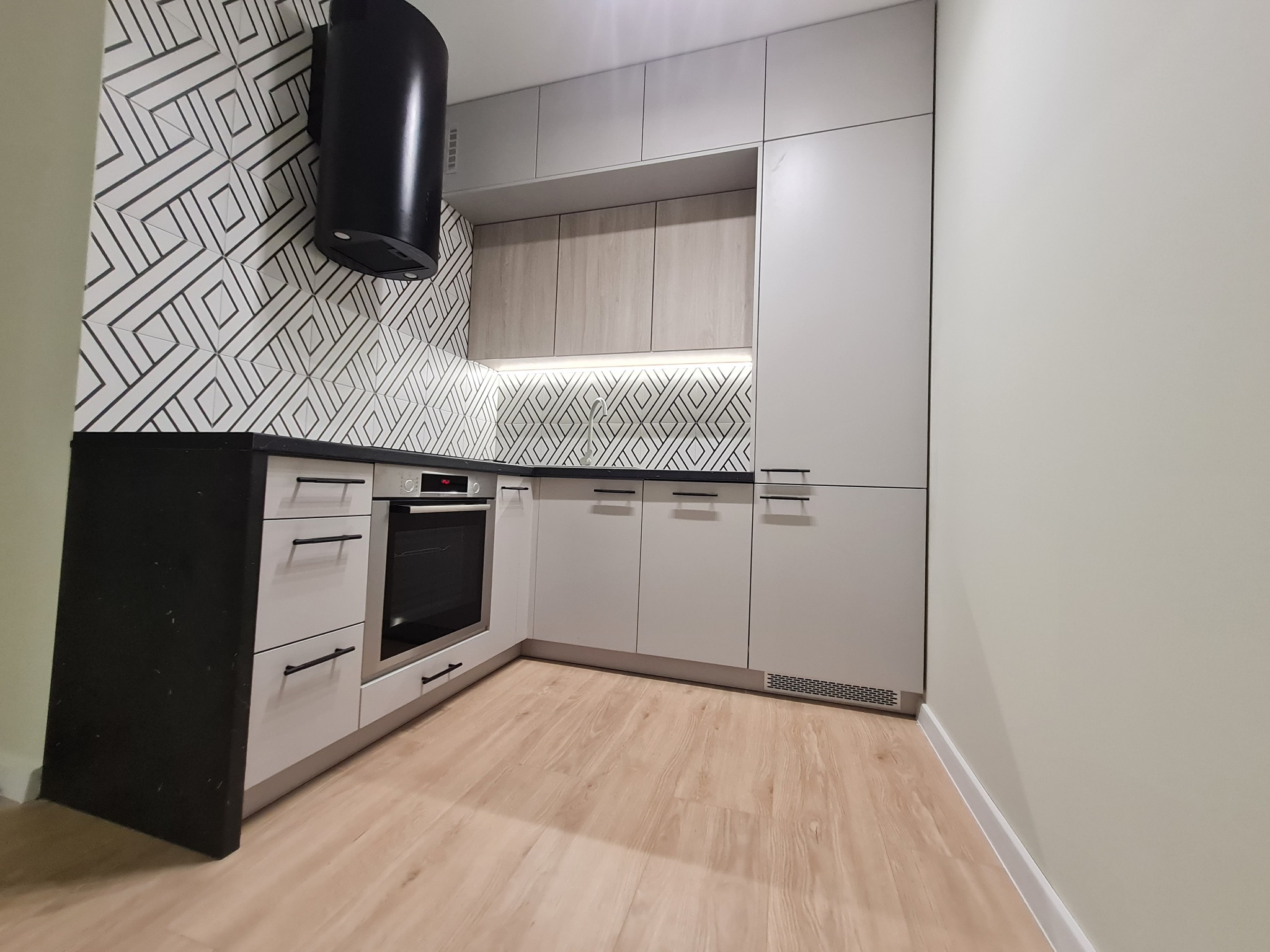 Nowoczesna kuchnia z białymi frontami, czarnym blatem i geometryczną tapetą. Uwagę przyciąga czarny okap i delikatne oświetlenie podszafkowe.