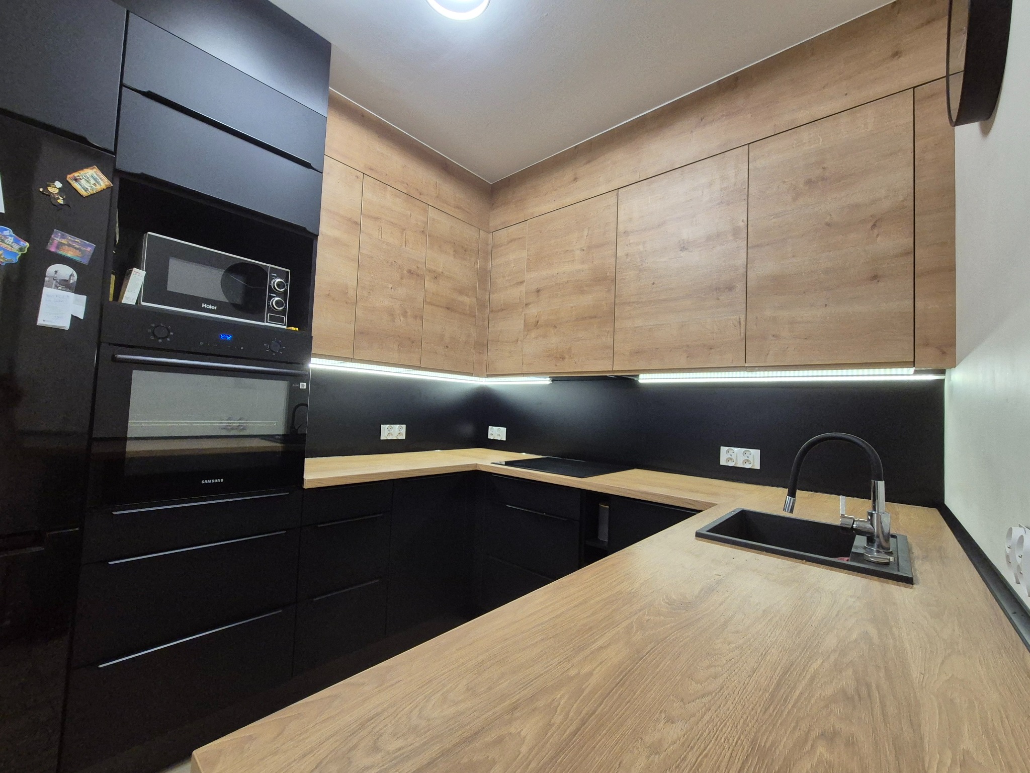 Nowoczesna kuchnia z drewnianymi szafkami górnymi i czarnymi dolnymi, oświetleniem LED podszafkowym i blatem imitującym drewno, z widocznym zlewem i baterią.