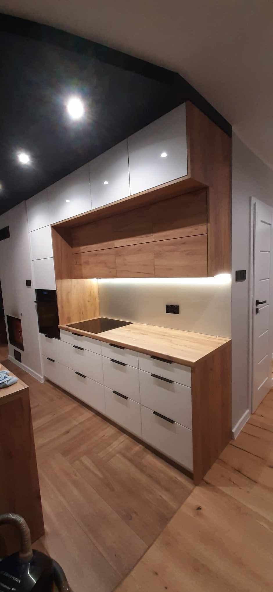 Nowoczesna kuchnia z białymi szafkami i drewnianymi akcentami, oświetleniem LED pod szafkami wiszącymi i czarnym sufitem. Parkiet w jodełkę na podłodze.