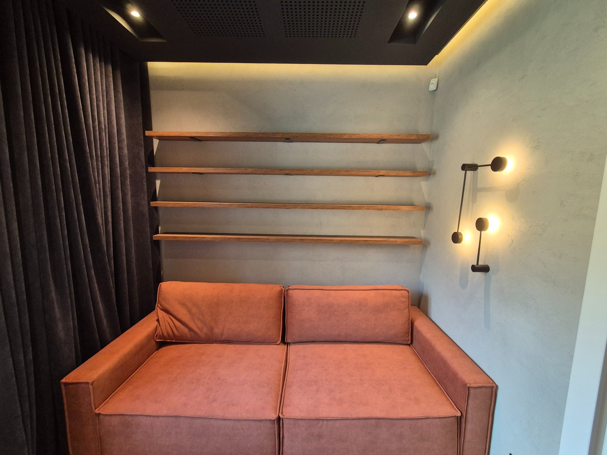 Minimalistyczny salon: brązowa sofa pod trzema drewnianymi półkami, oświetlona kinkietem i listwą LED, z zasłoną po lewej. Nowoczesny design.
