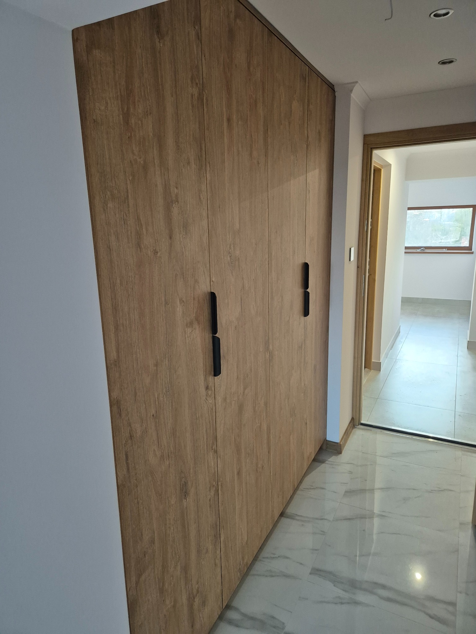 Wysoka, zabudowana szafa z drewnianymi frontami i czarnymi uchwytami, wkomponowana we wnętrze mieszkania z marmurową podłogą.