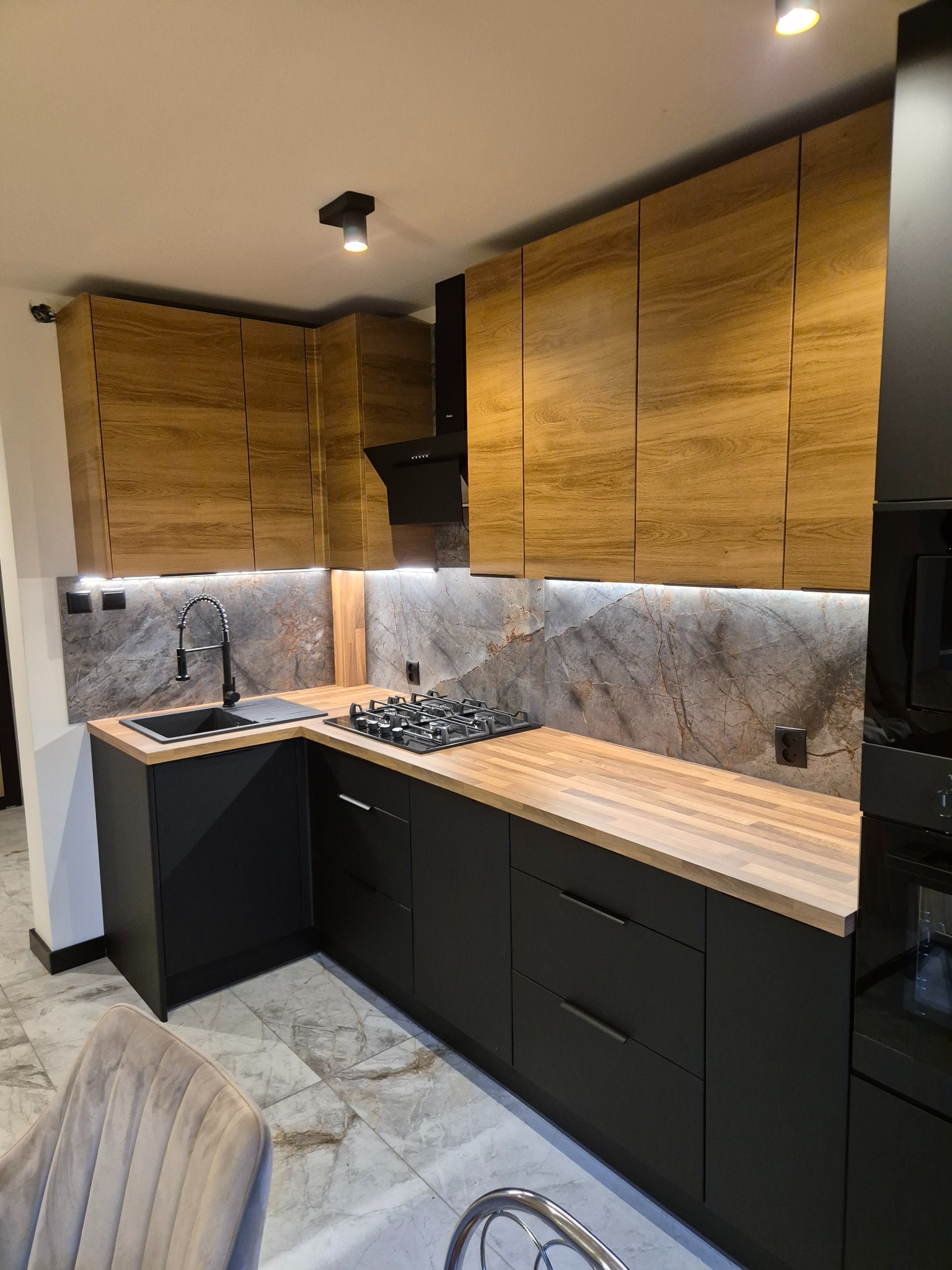 Nowoczesna kuchnia z drewnianymi szafkami górnymi i czarnymi dolnymi, oświetleniem LED pod szafkami i marmurowym backsplash'em. Widoczna płyta gazowa i czarny zlew.