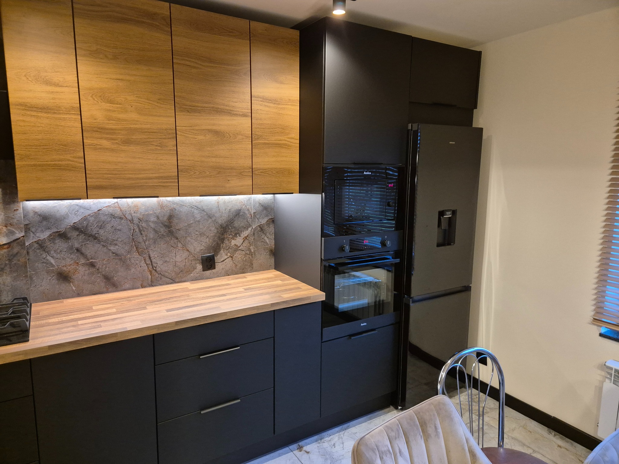 Nowoczesna kuchnia z drewnianymi szafkami górnymi i czarnymi dolnymi, marmurowym backsplashiem i drewnianym blatem. Widoczny sprzęt AGD.