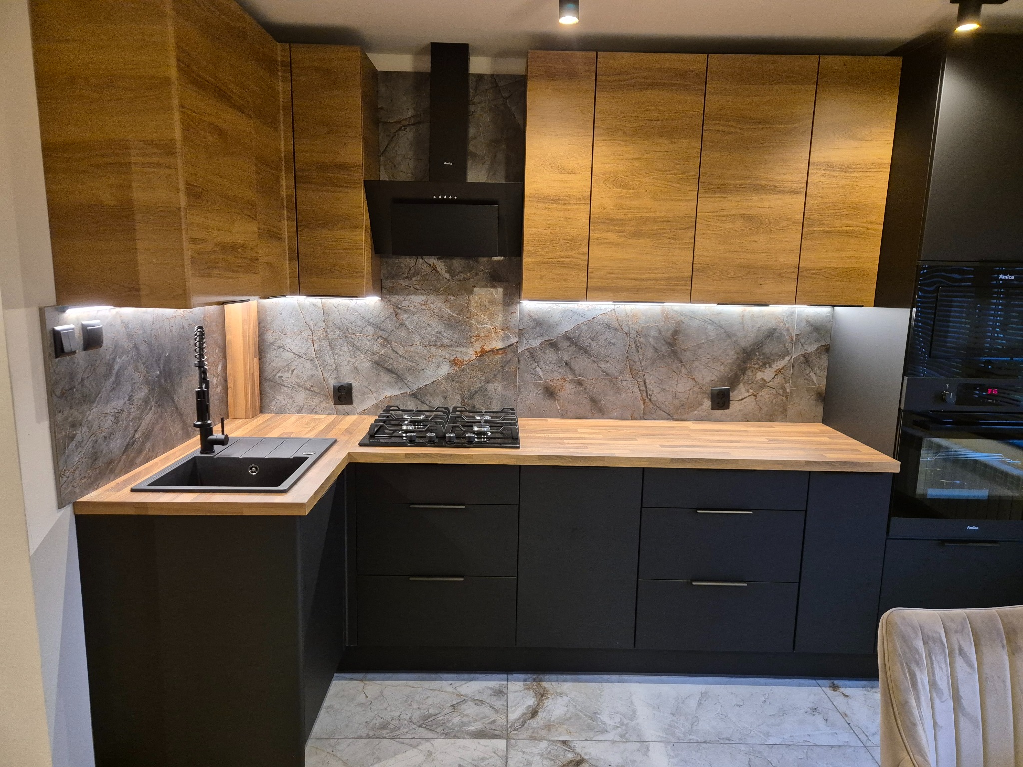 Nowoczesna kuchnia z drewnianymi szafkami górnymi i czarnymi dolnymi, blatem z jasnego drewna i marmurową ścianą. Widoczny zlew, płyta gazowa i piekarnik.