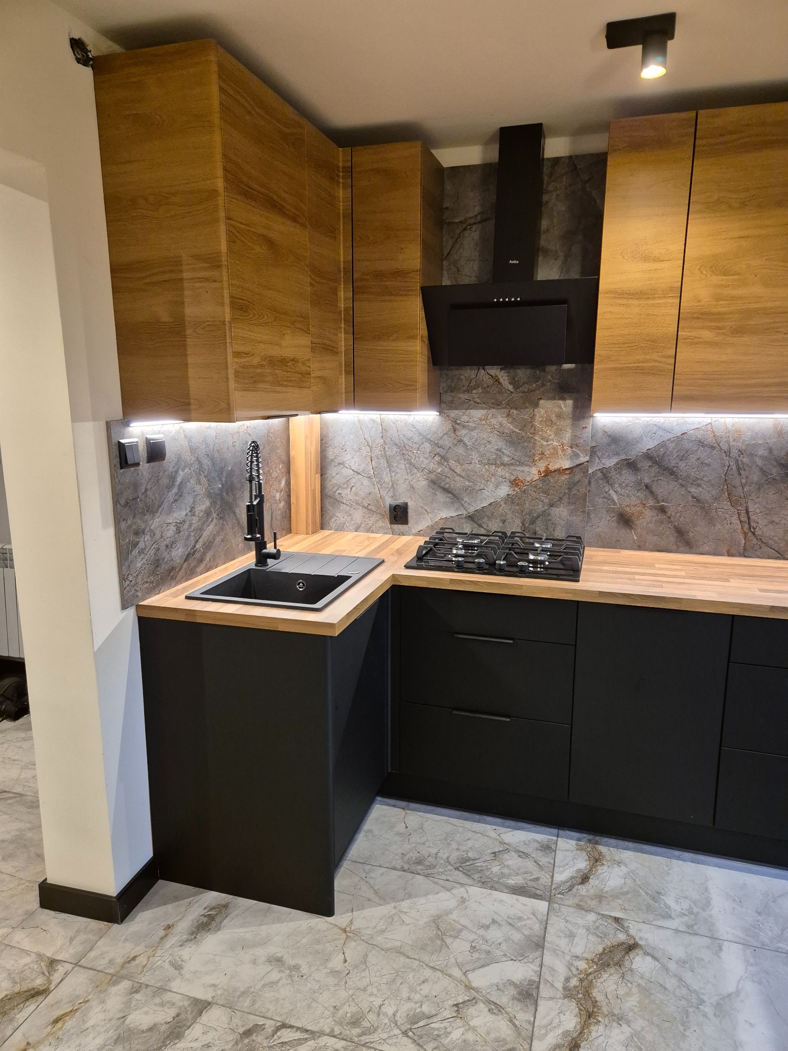Nowoczesna kuchnia z drewnianymi szafkami górnymi i czarnymi dolnymi, marmurowym blatem i płytkami. Oświetlenie LED podkreśla detale. Widoczny fragment płyty gazowej.