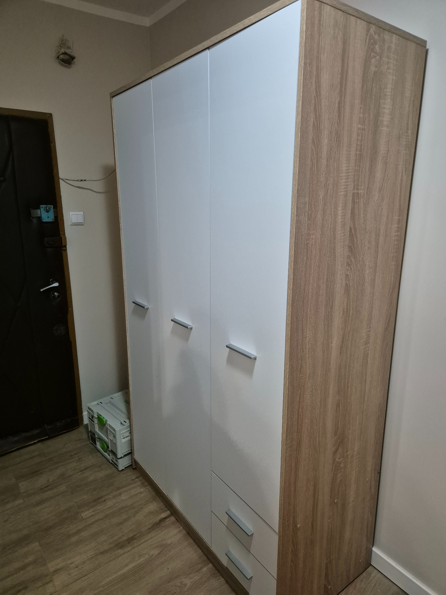 Nowoczesna szafa trzydrzwiowa z białymi frontami i drewnopodobnymi bokami, z szufladami na dole, stojąca w rogu pokoju obok drzwi.